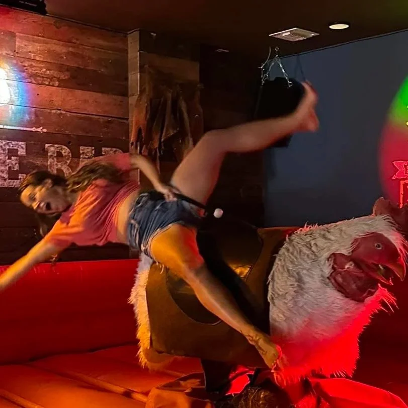 Person riding a large, realistic  mechanical bucking  ROOSTER ride , inside Saddle Bar  with wooden walls and neon lighting.
