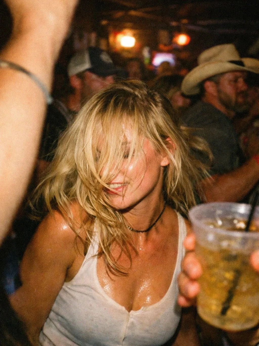 Woman with wet hair smiling and holding a drink at a crowded bar or party.