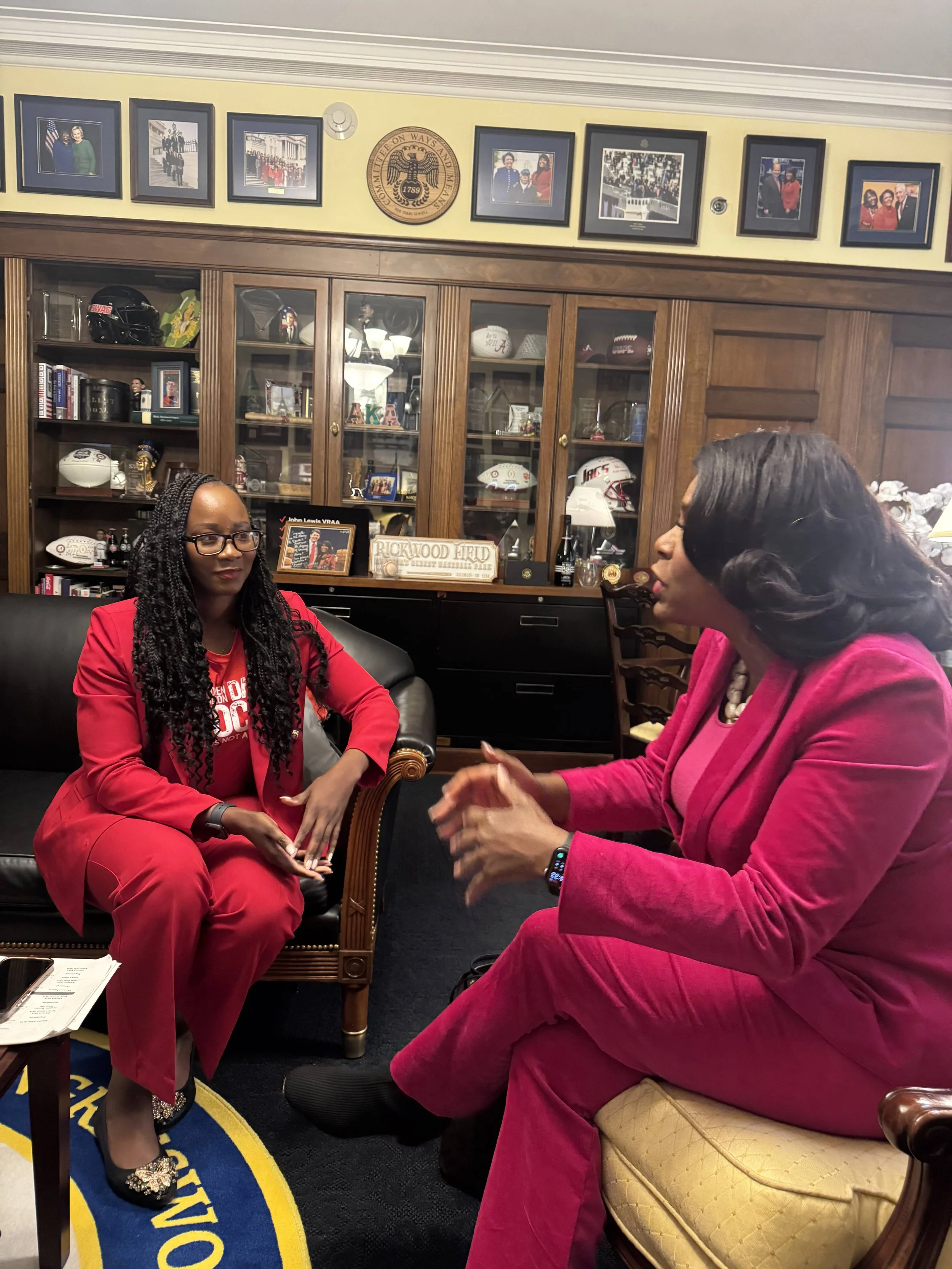 Two women in pink suits having a conversation in an office with wood display cabinets and framed photographs on the wall.