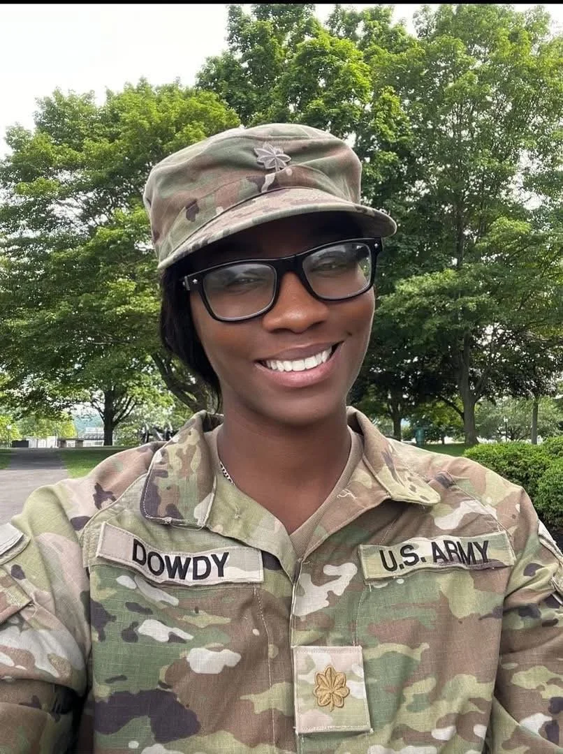 A woman in U.S. Army camouflage uniform and cap, wearing glasses, smiling outdoors with green trees in the background.