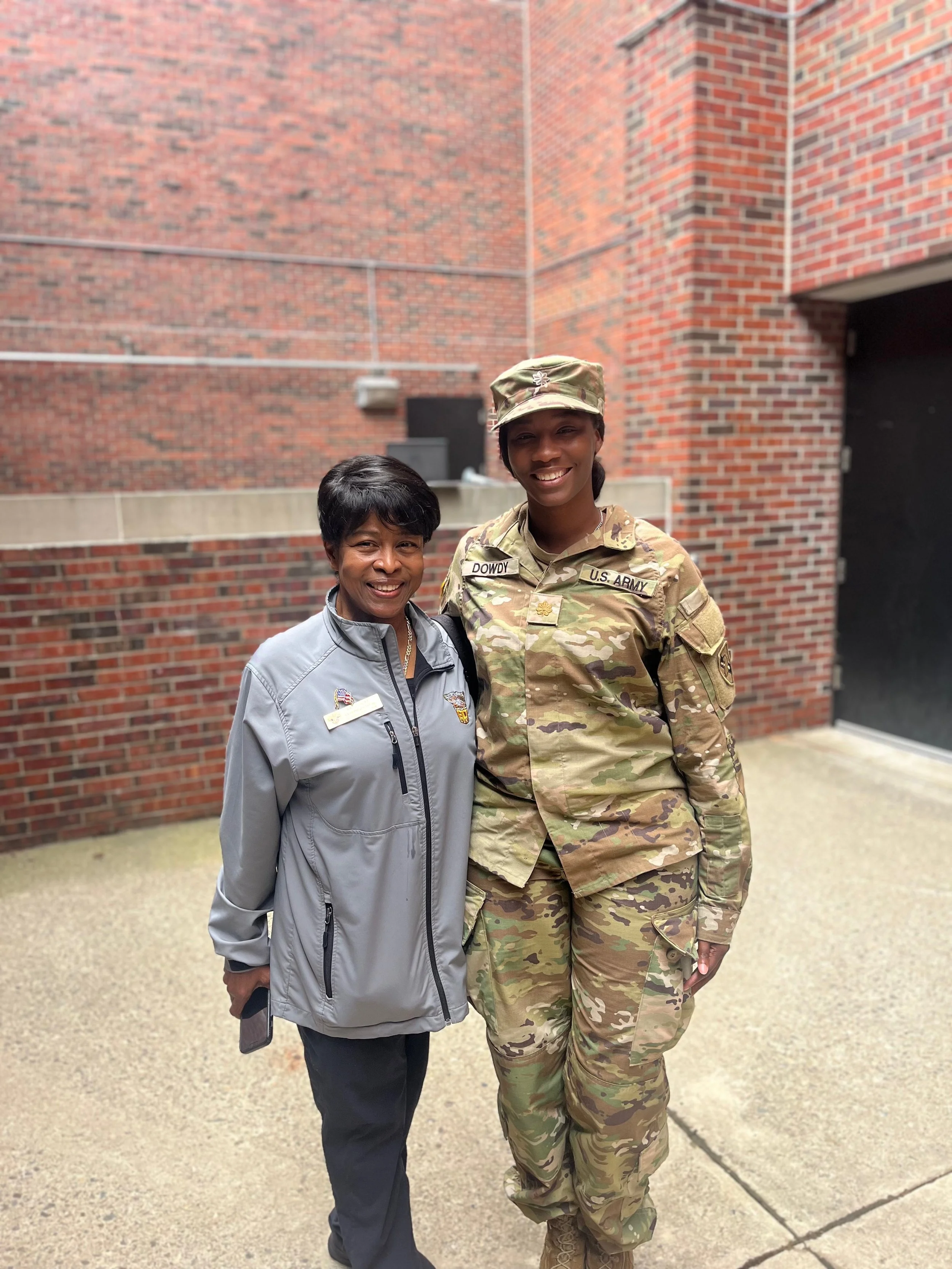 A woman and a female U.S. Army soldier standing outdoors in front of a brick building, smiling and posing together.