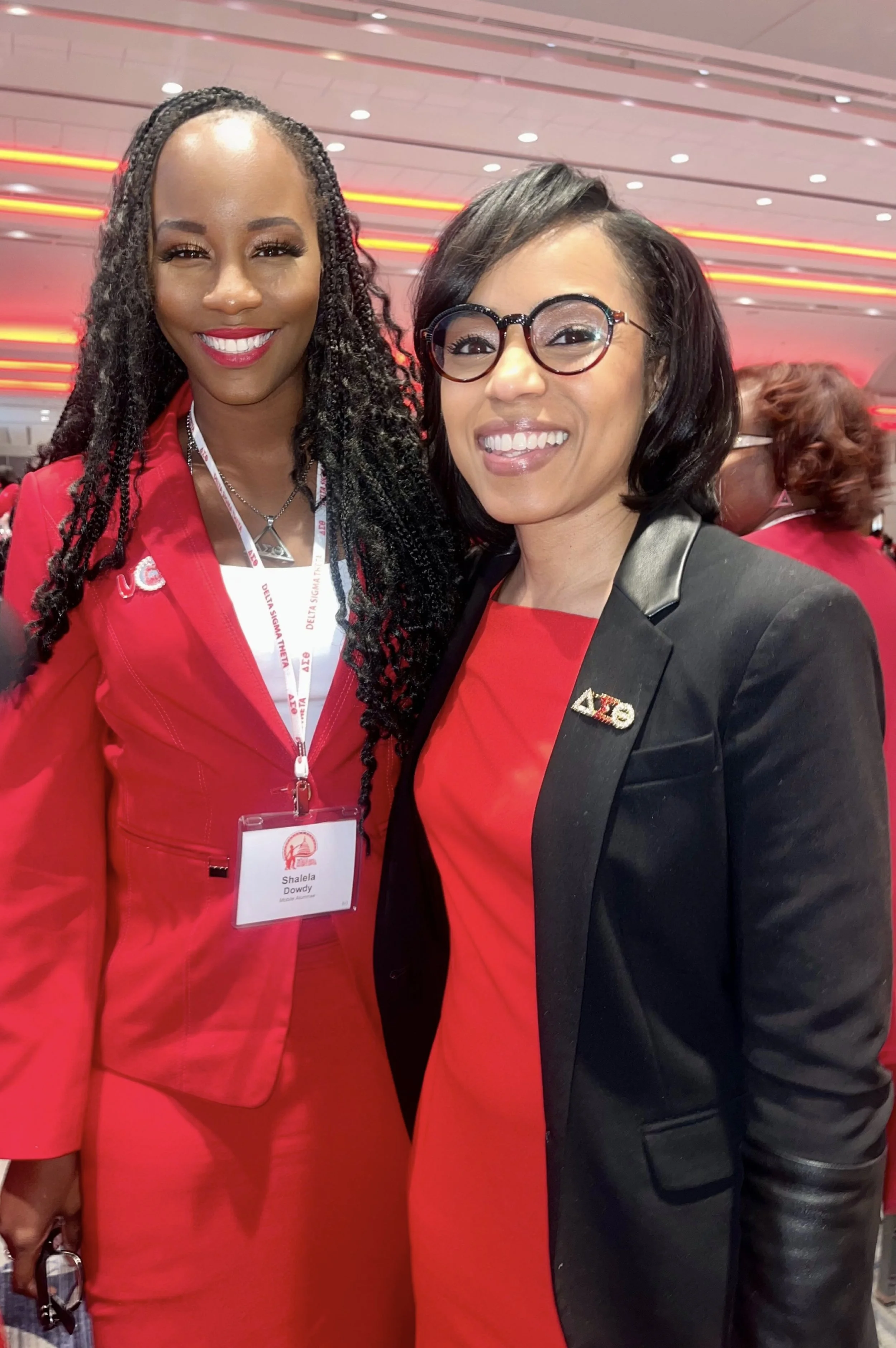 Two women smiling at a conference, wearing red and black outfits. The woman on the left has long braided hair and a red suit with a name tag. The woman on the right has shoulder-length black hair, glasses, a black blazer with a Delta Sigma Theta soro