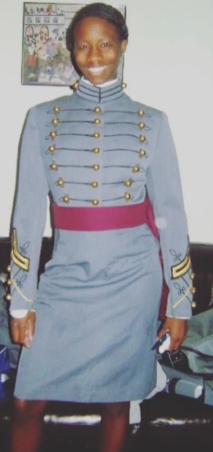 A woman dressed in a gray military-style uniform with gold buttons and embroidery, standing indoors with a small smile, holding a phone and keys, and a colorful picture on the wall behind her.