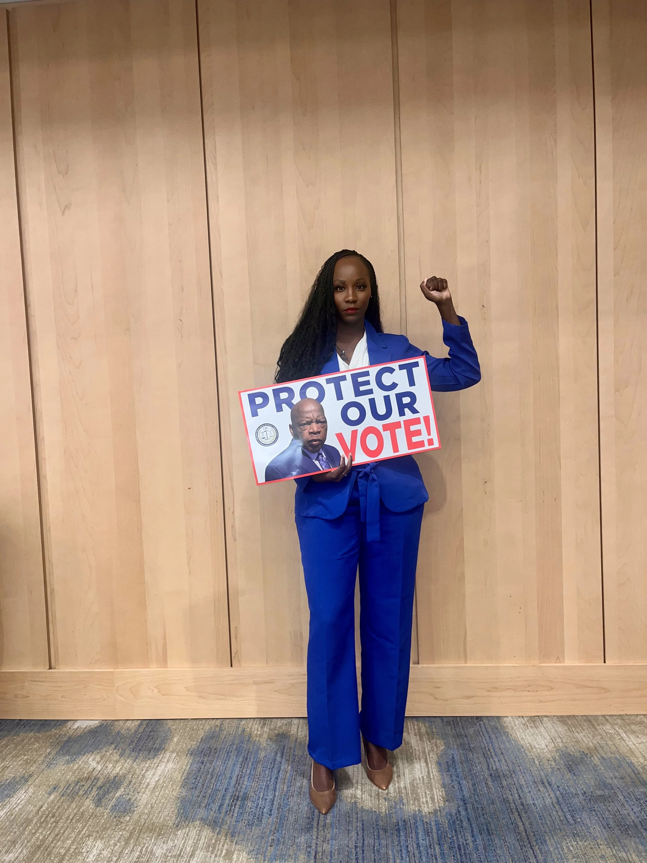 A woman standing indoors against a wooden wall, wearing a blue pantsuit and beige high heels, holding a political sign that says 'PROTECT OUR VOTE!' with an image of a man in a suit and a seal of a government agency.
