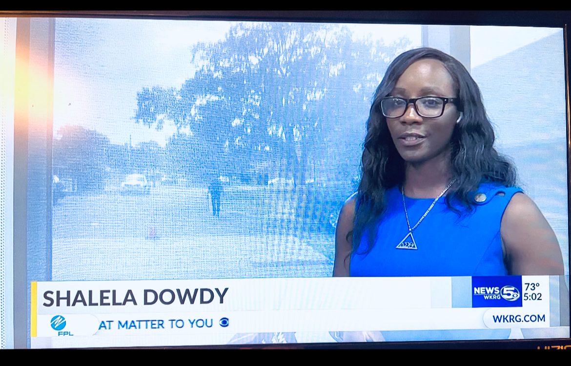 A female news reporter with glasses and wavy black hair, wearing a blue top and a triangular pendant necklace, standing outdoors in front of a window showing a tree and street scene.