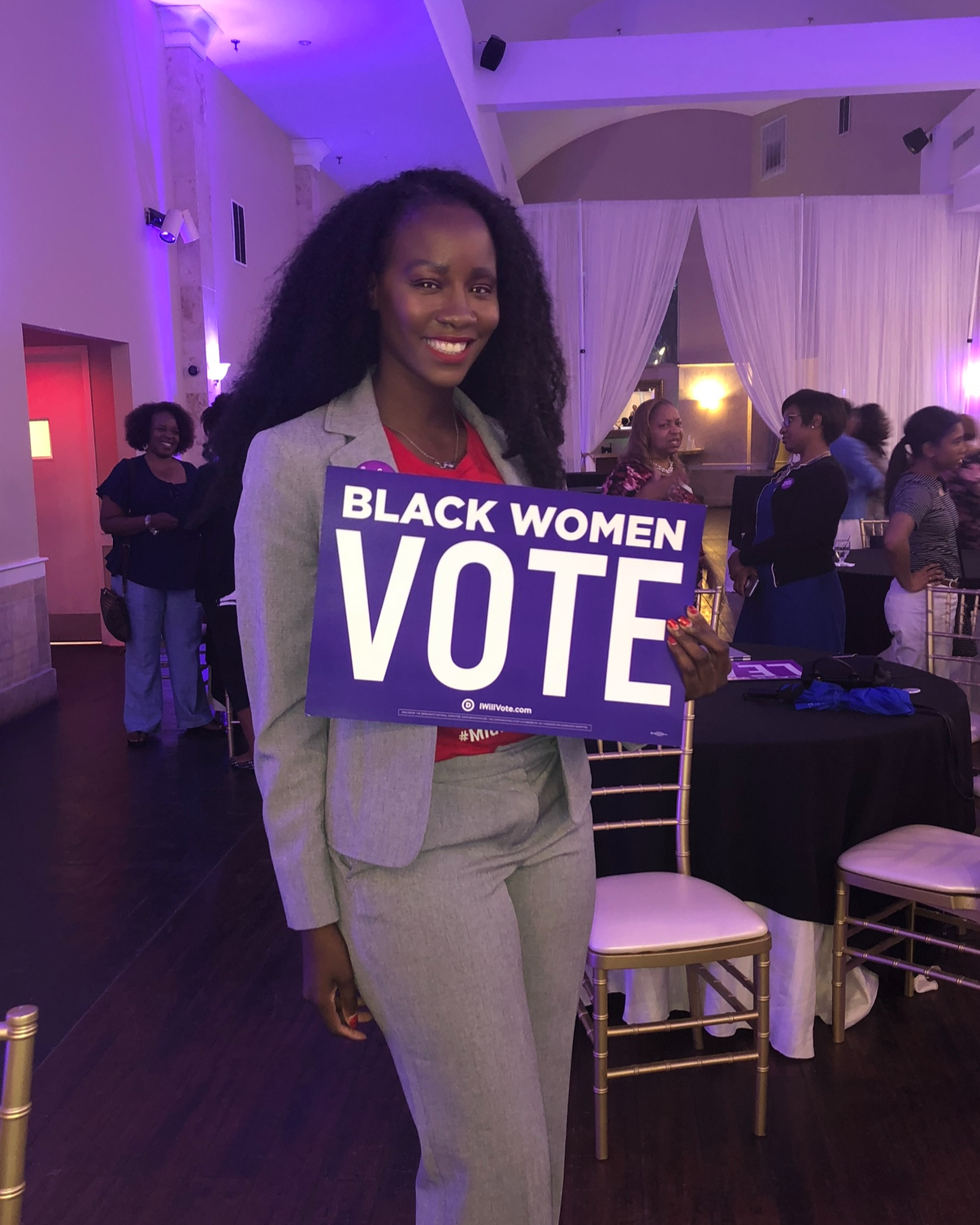 A woman in a gray suit holding a purple sign that says "Black Women Vote" at an indoor event with purple lighting and several people in the background.