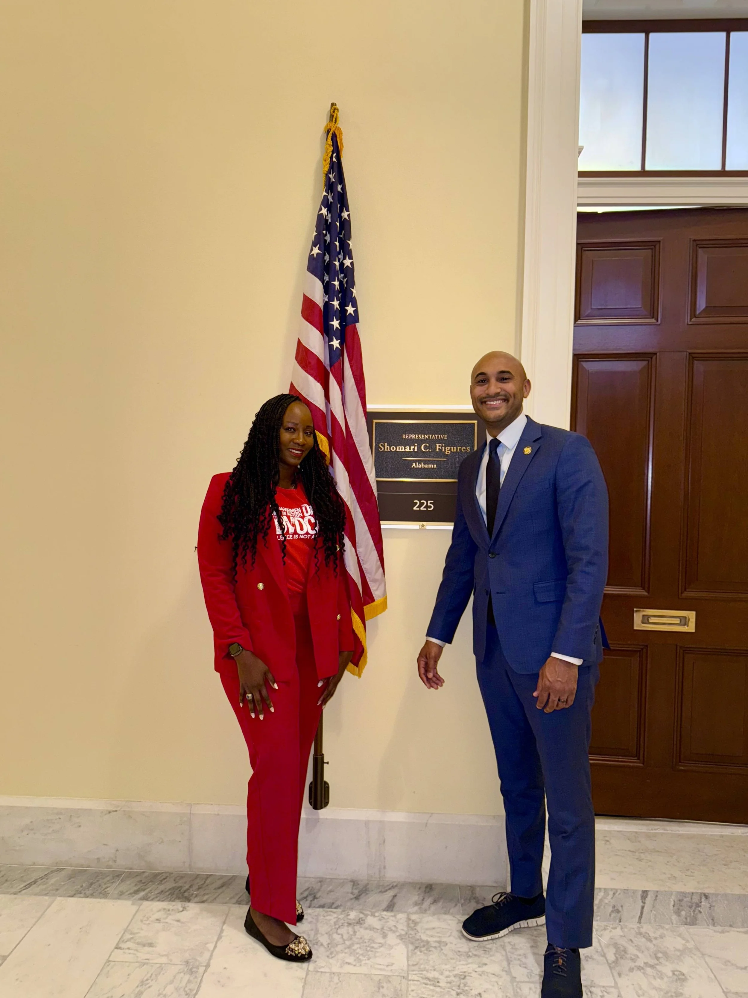 Two individuals, a woman and a man, standing in front of an American flag and a plaque indicating the office of Shomari C. Figures, a representative from Alabama, in a government building.