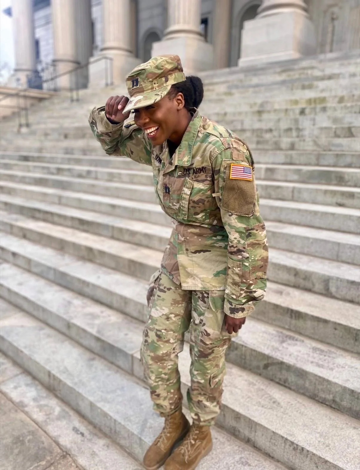 A smiling woman in U.S. Army camouflage uniform stands on stairs outside a government building, adjusting her hat.
