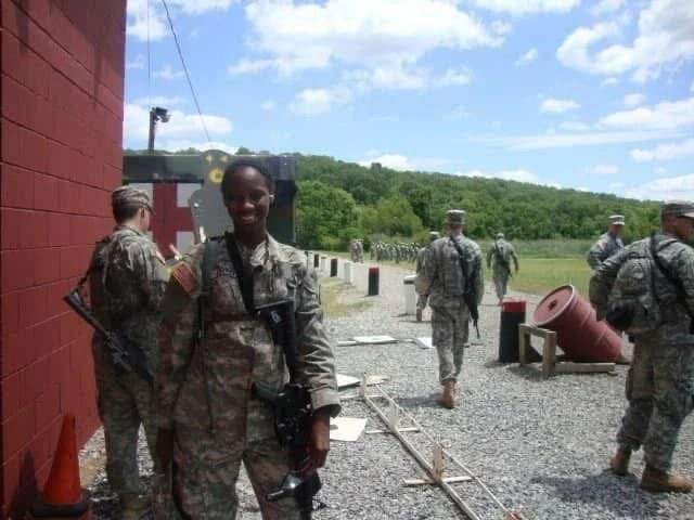 A young woman in military uniform smiling, standing outdoors with several other soldiers in the background near a red brick building on a gravel surface under a blue sky with scattered clouds.