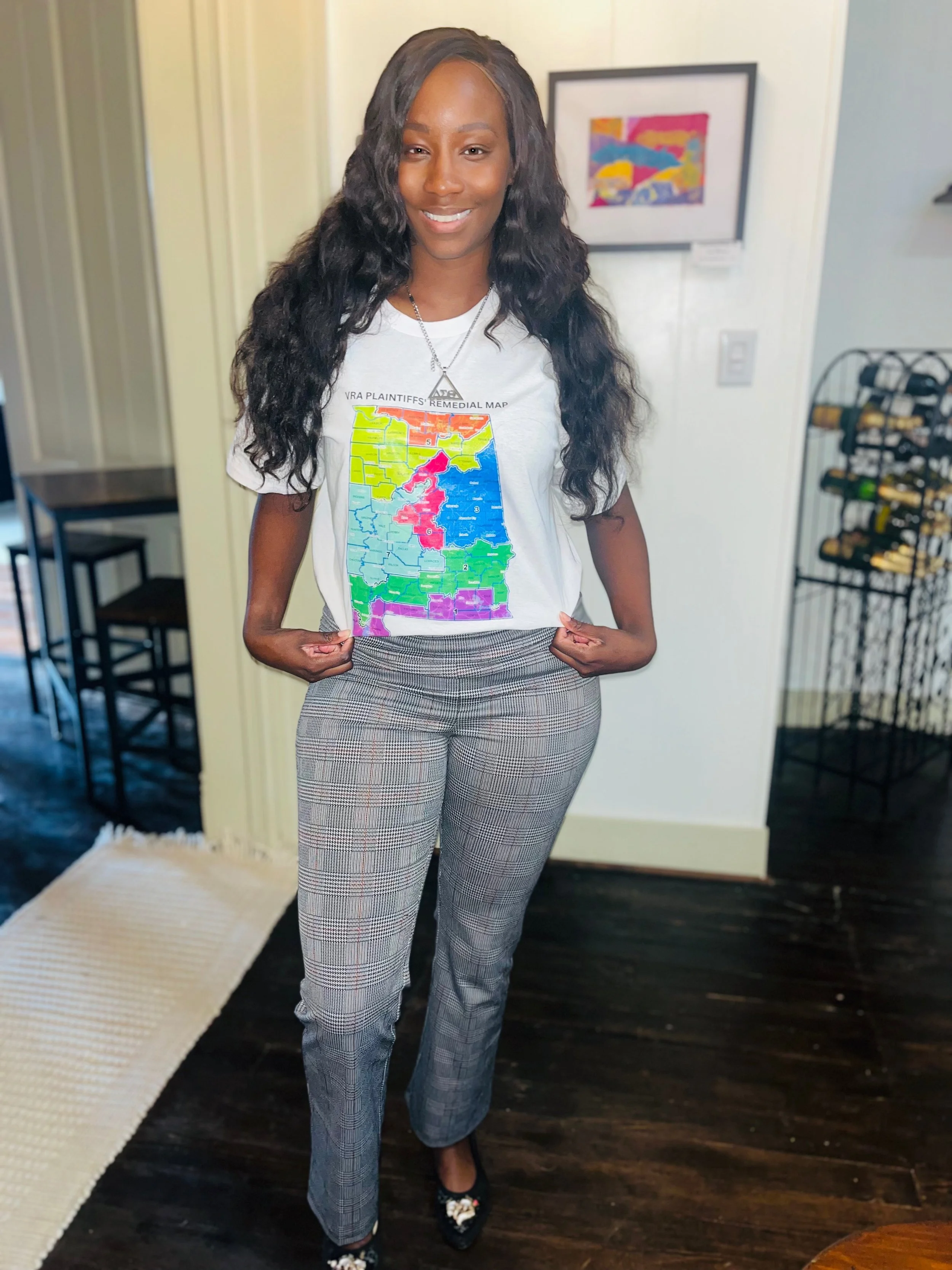 Woman with long curly hair standing indoors, wearing a white T-shirt with a colorful map of Arkansas, plaid pants, and black shoes.