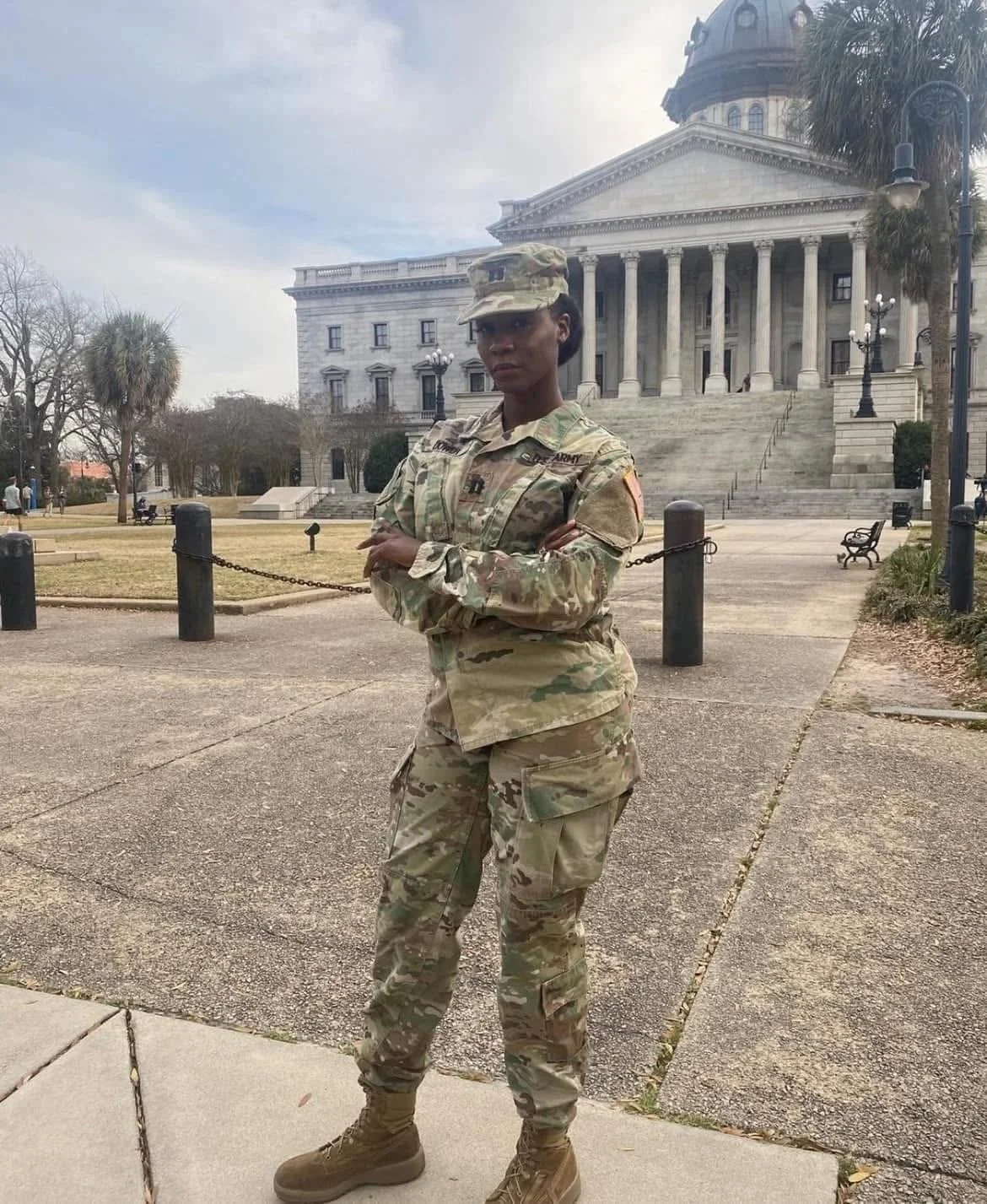 A female soldier in camouflage uniform standing with arms crossed in front of a historic government building with columns and a dome.