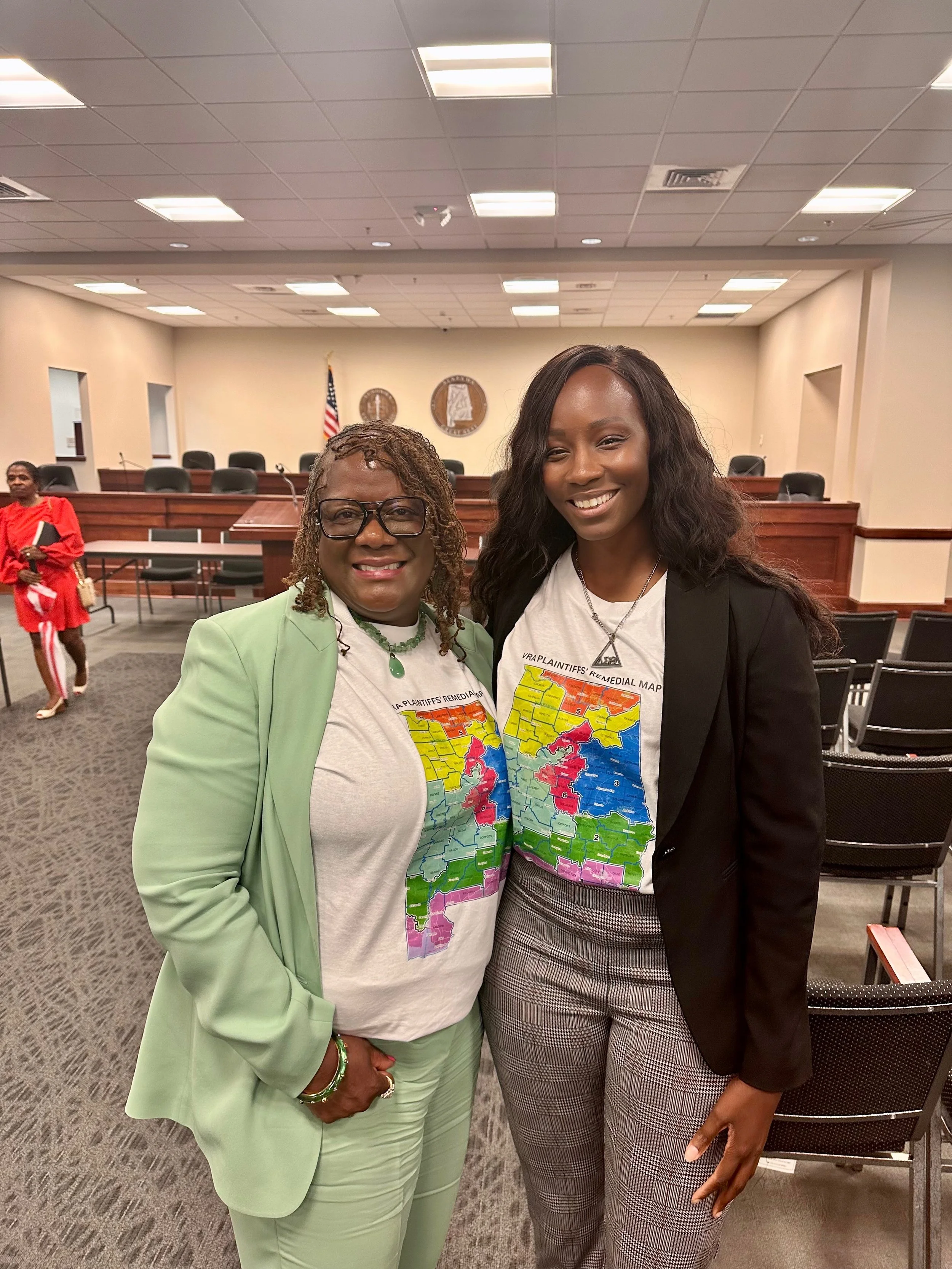 Two women standing inside a courtroom or government building, wearing white T-shirts with a colorful map of various states and the text 'VRA PLAINTIFFS' REMEDIAL MAP'. One woman is wearing a light green suit, glasses, and jewelry, while the other is 