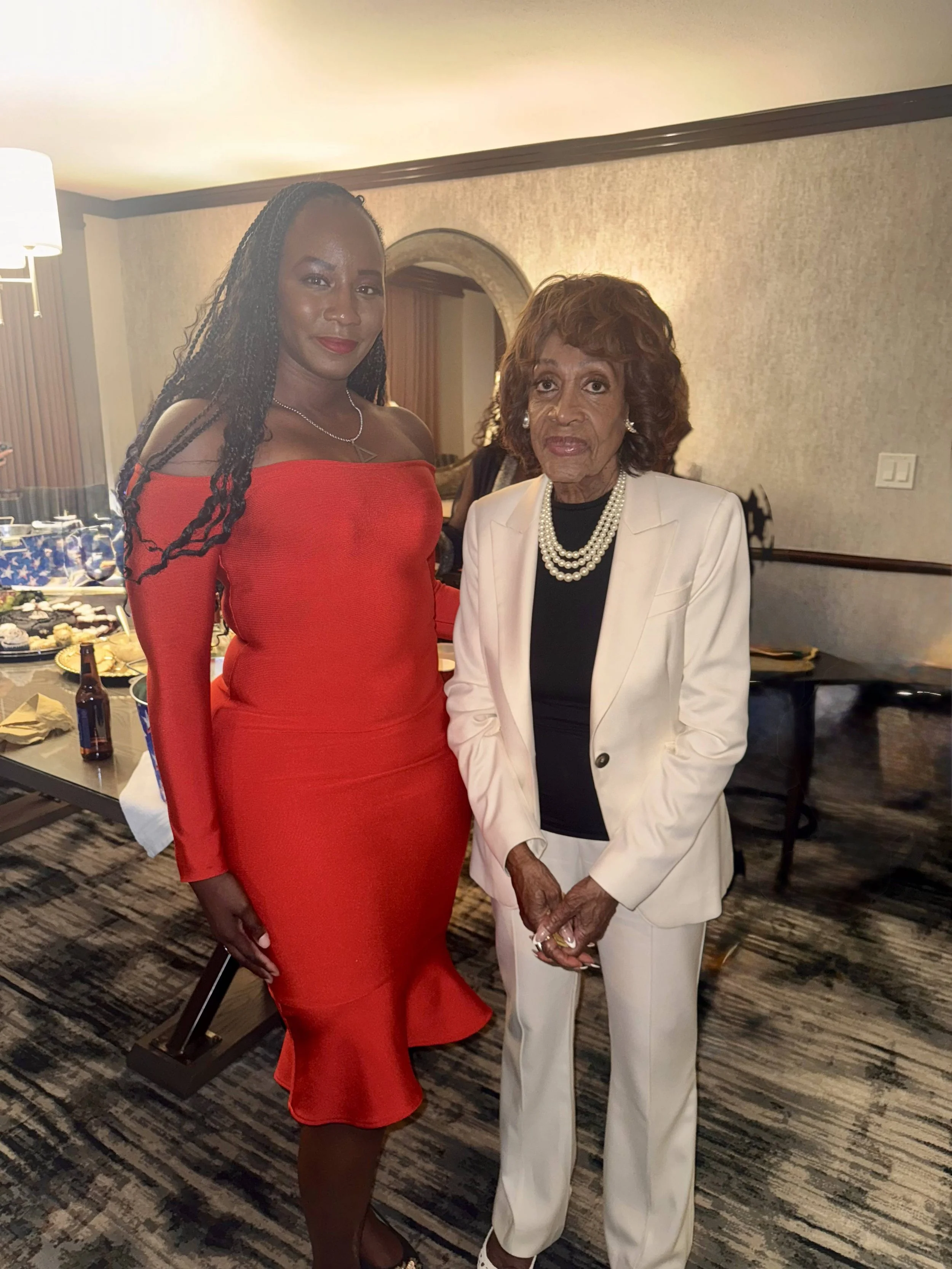 Two women pose together at an indoor event. The woman on the left is wearing a red off-shoulder dress and the woman on the right is dressed in a white suit with a black top, pearl necklace, and earrings.