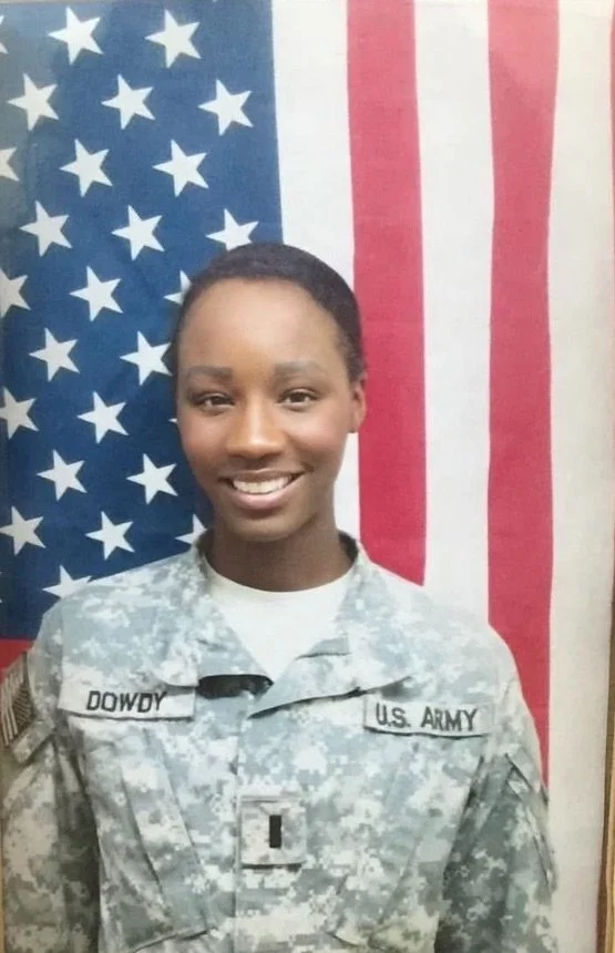 A woman in U.S. Army camouflage uniform standing in front of an American flag.