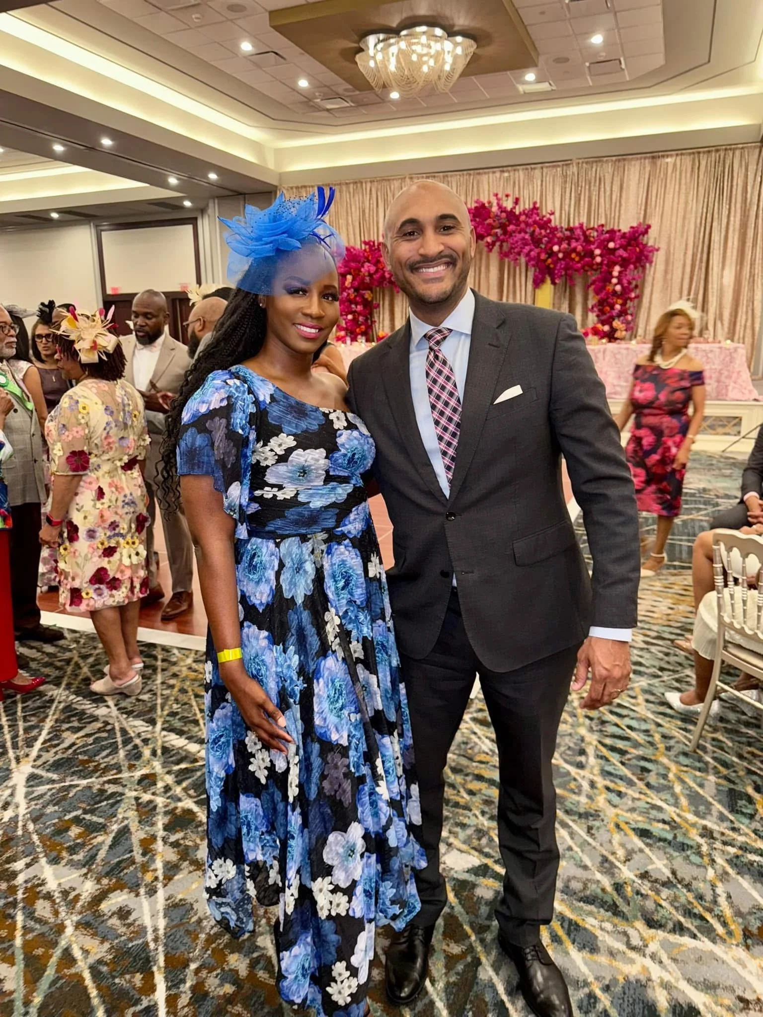 A smiling couple at a formal event, the woman dressed in a blue floral gown with a matching blue fascinator, and the man in a dark suit with a patterned tie. They are standing in a decorated banquet hall with other guests in colorful attire.