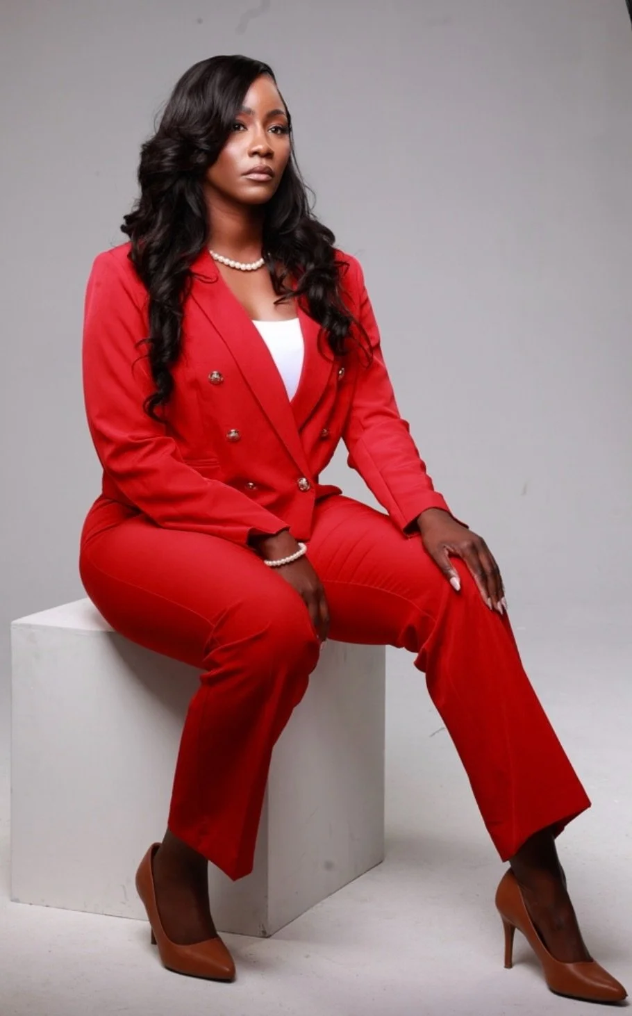 Shalea Dowdy sits on a white cube wearing a red pantsuit, white top, pearl necklace, pearl bracelet, and tan high heels, with long wavy hair and a confident expression against a plain gray background.
