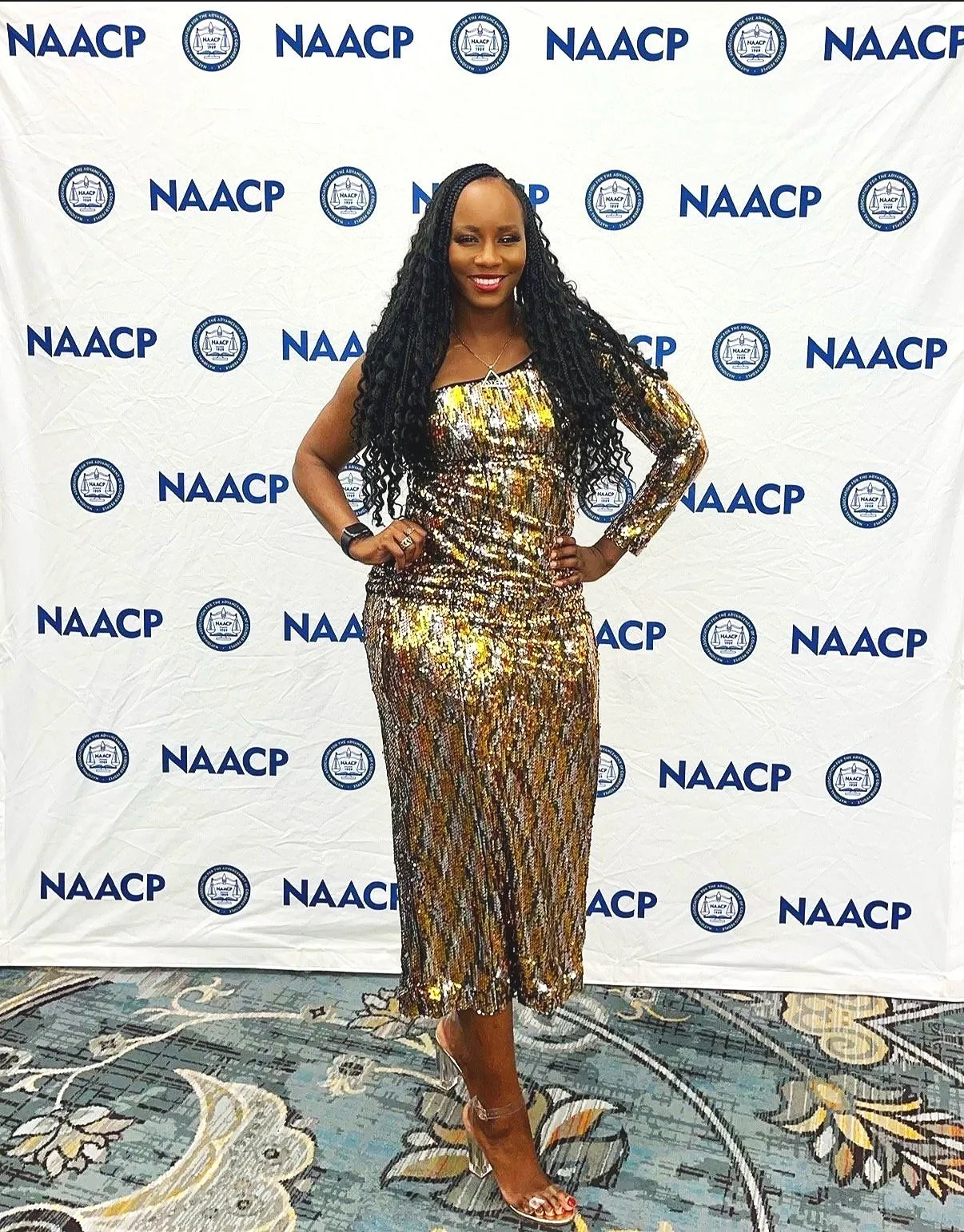 Woman in a shiny, patterned dress standing in front of a NAACP backdrop at an event