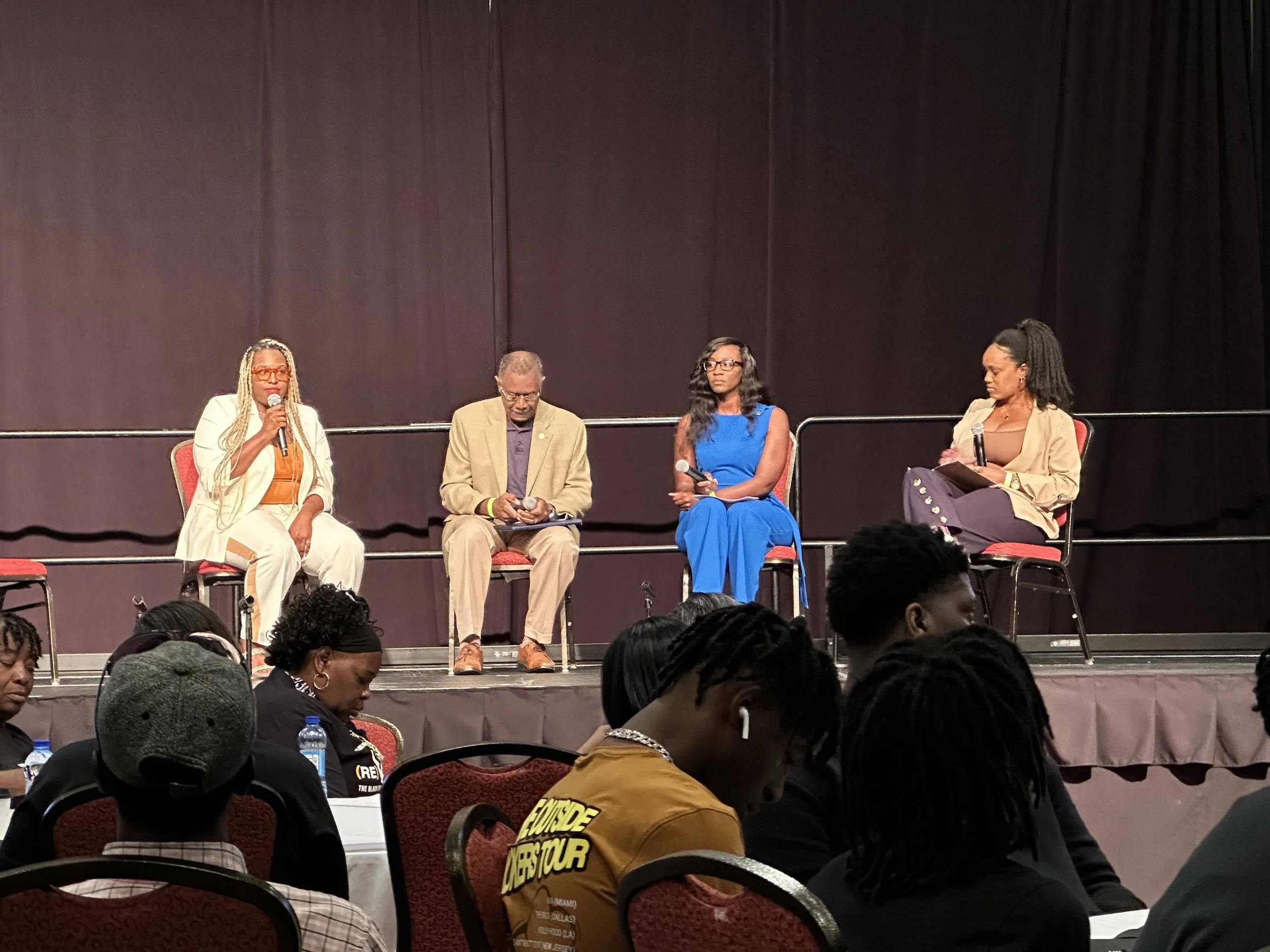 Four panelists sitting on stage during a panel discussion, with an audience in front of them. The panelists include three women and one man, with one woman speaking into a microphone. The stage has a dark backdrop and red chairs, and the audience con