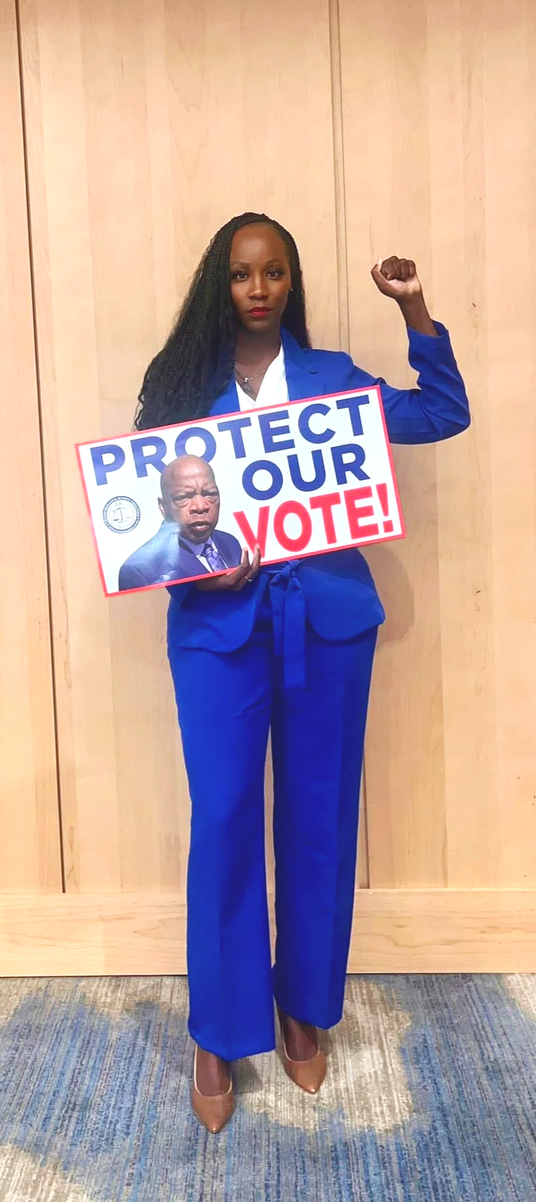 A woman in a blue pantsuit holding a sign that says 'Protect Our Vote!' with a picture of a man pointing a finger.