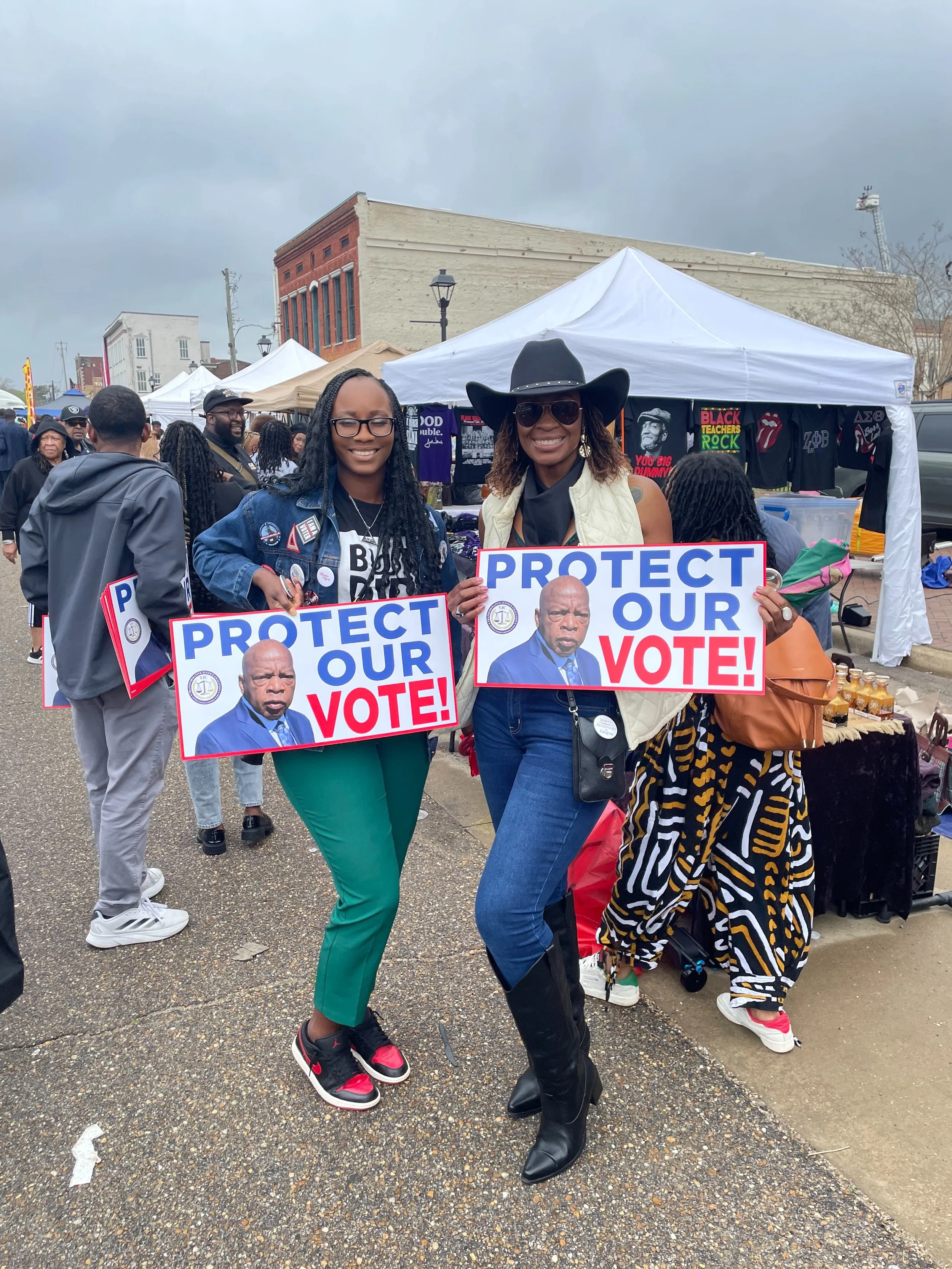 Two women holding signs that say "Protect Our Vote!" at a crowded outdoor event or protest.