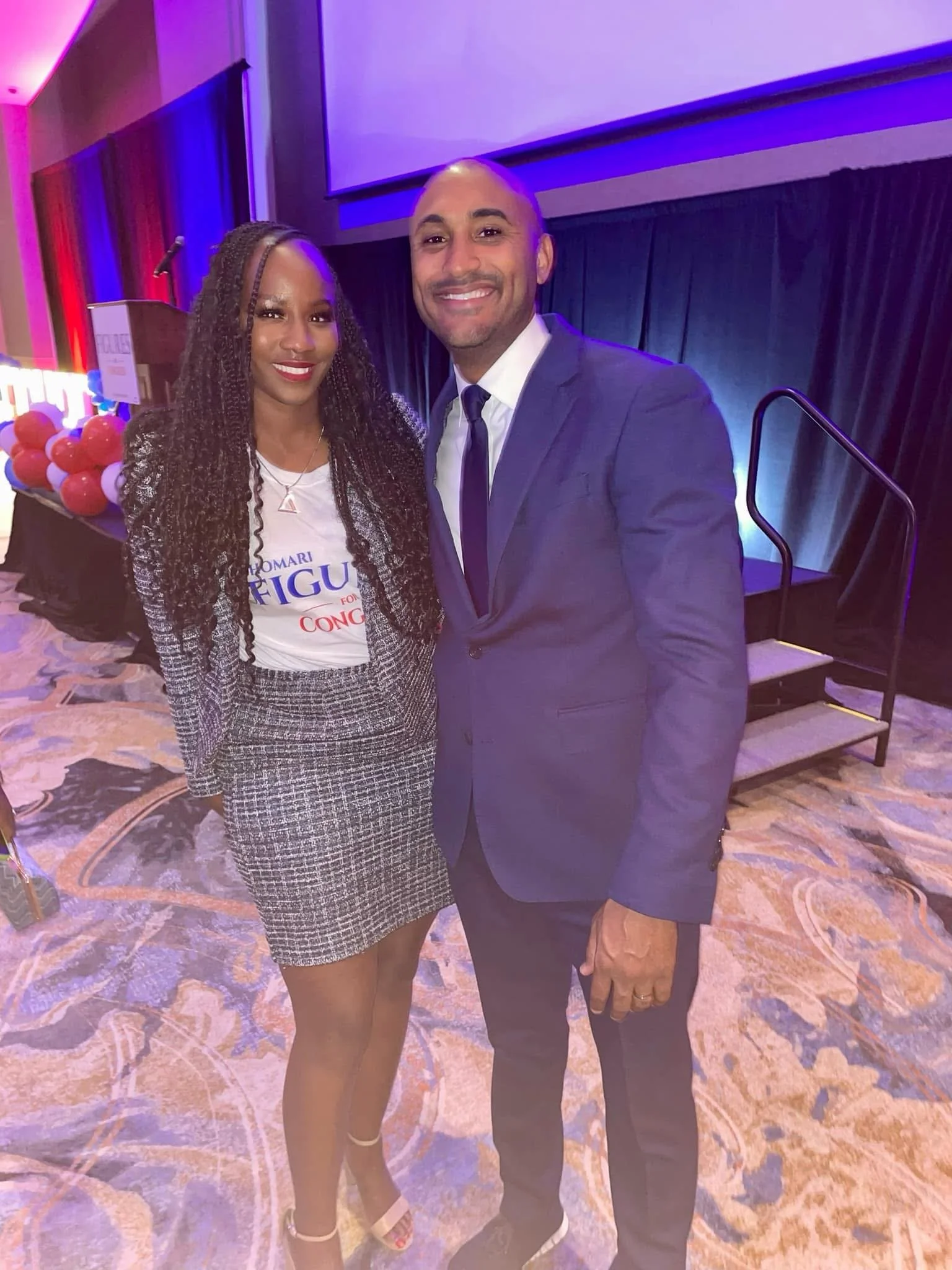 A woman and a man are standing together at a formal event. The woman has long curly hair and is wearing a plaid skirt and blazer, a white t-shirt, and high heels. The man is wearing a blue suit, white shirt, and dark tie. They are smiling at the came