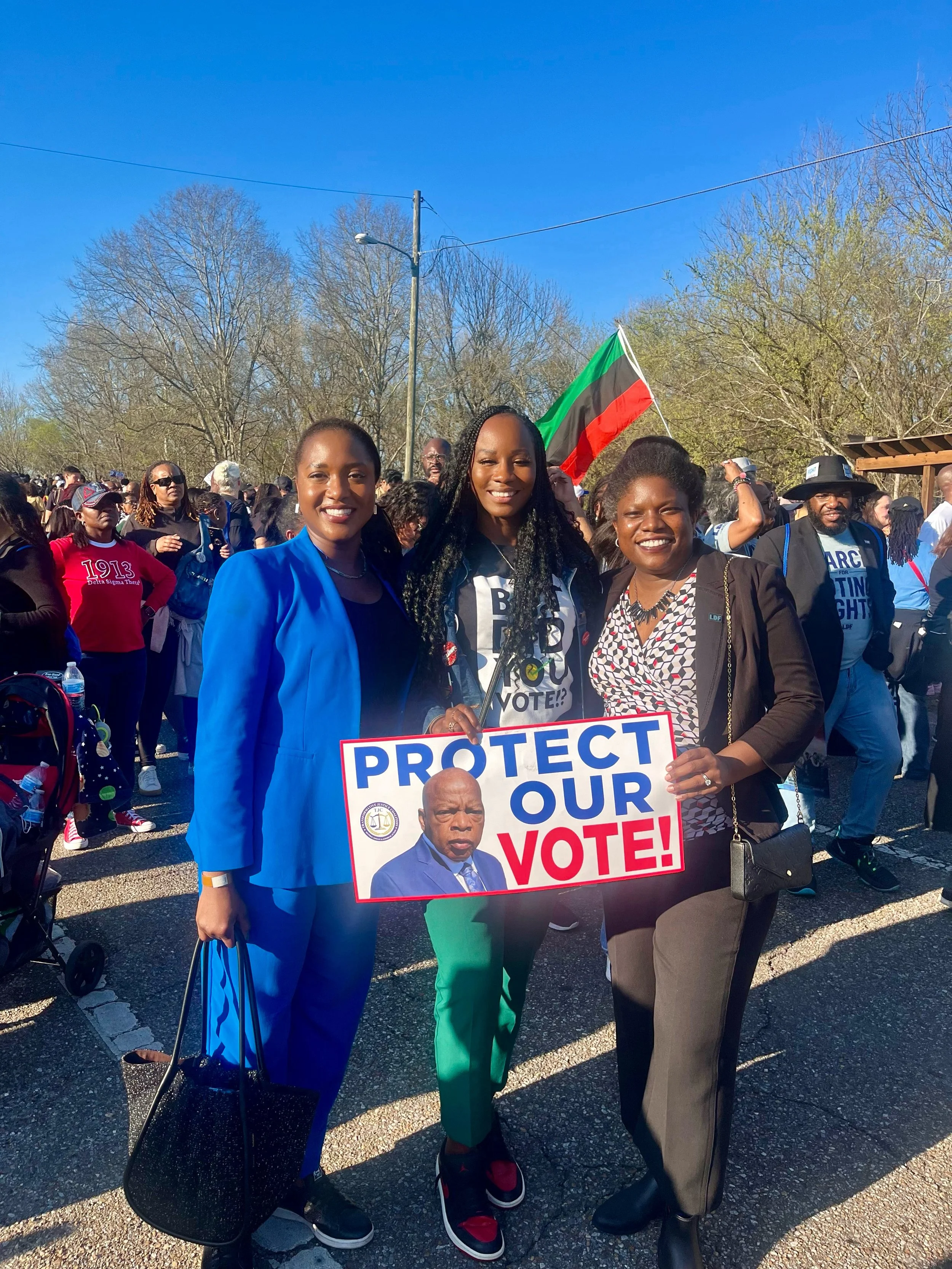 Three women at a rally holding a sign that says 'Protect Our Vote!' with an image of a man in a suit, and a person in the background waving a flag.