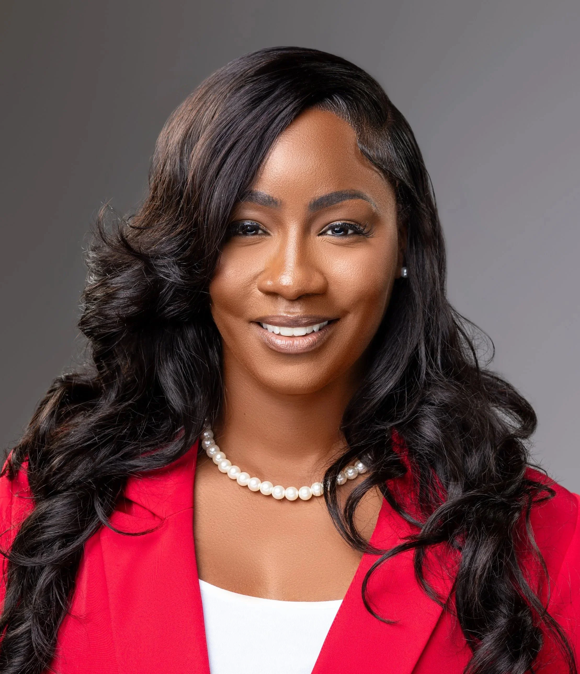 Shalea Dowdy  woman with long wavy black hair wearing a red blazer, white top, pearl necklace, and pearl earrings against a gray background.