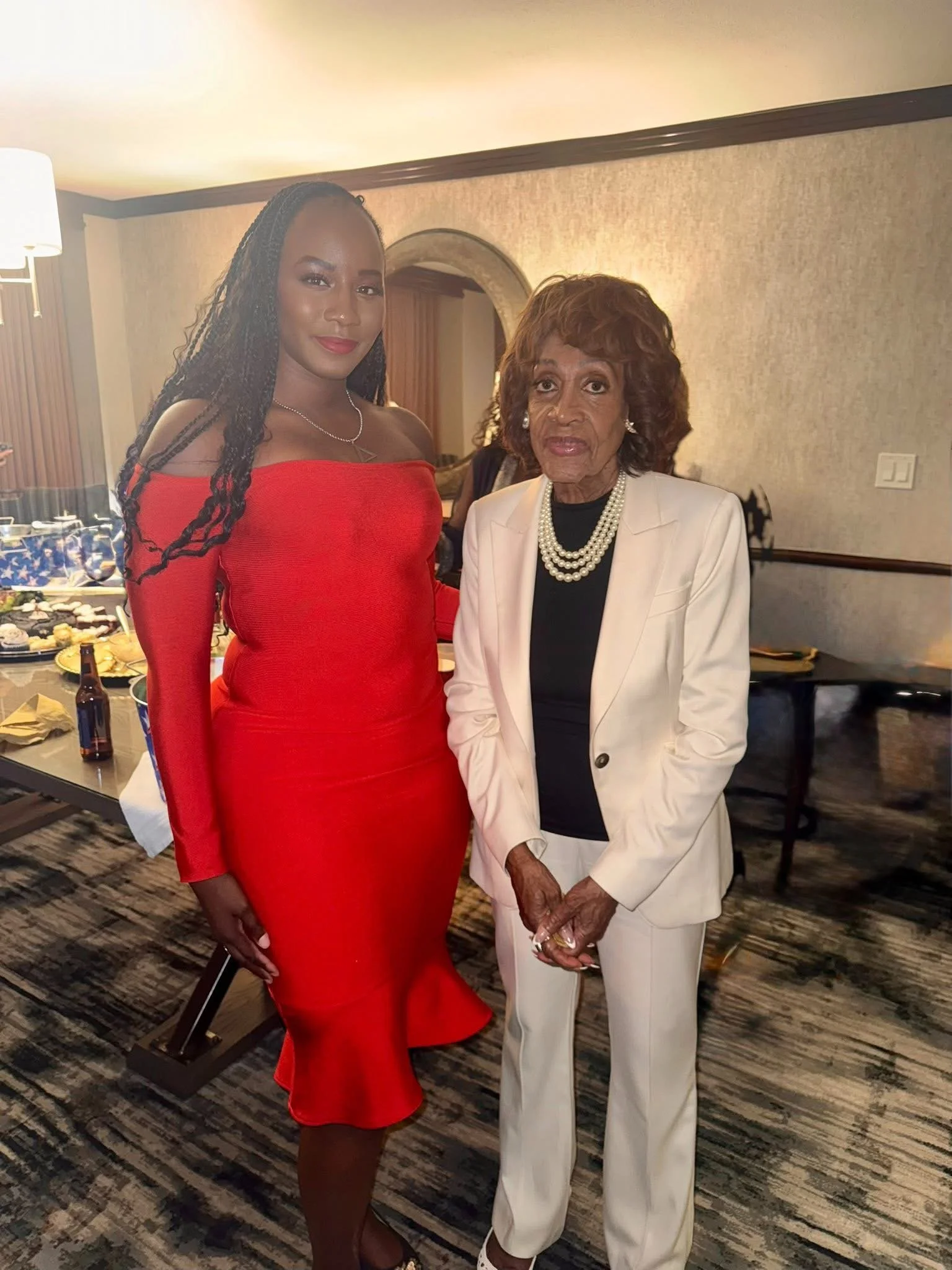 Two women standing together at a social gathering, with a table of snacks and drinks in the background.