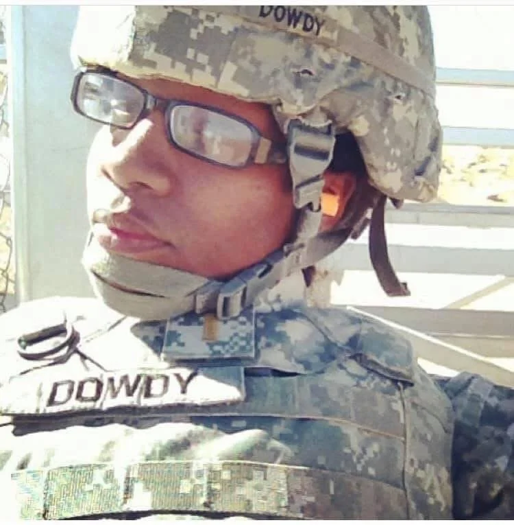 Close-up of a soldier in camouflage uniform with glasses and helmet, outdoors with bright sunlight.