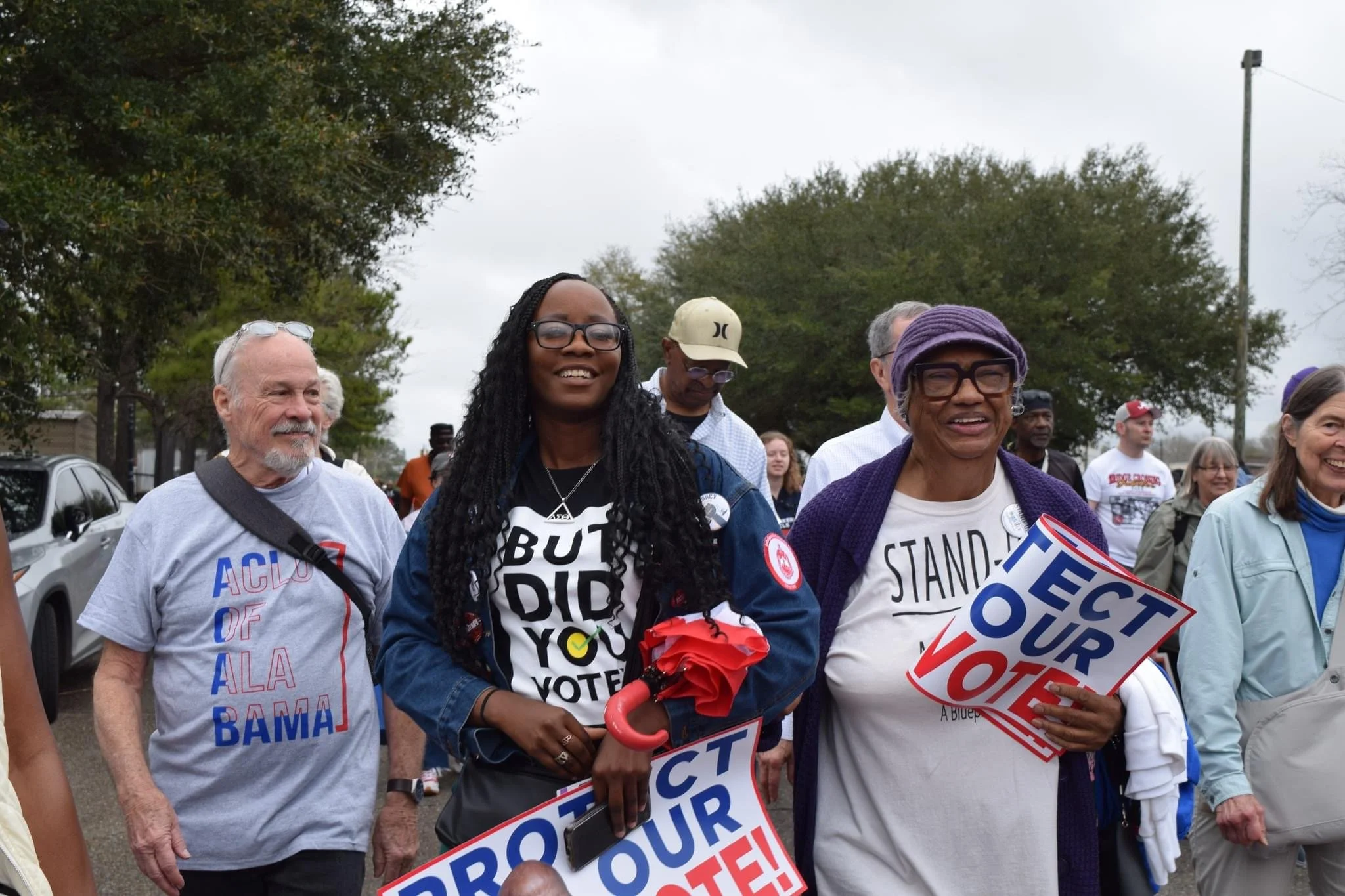 Group of diverse people walking outdoors during a protest, holding signs that read "Protect Our Vote" and wearing clothing supporting voting rights and political causes.