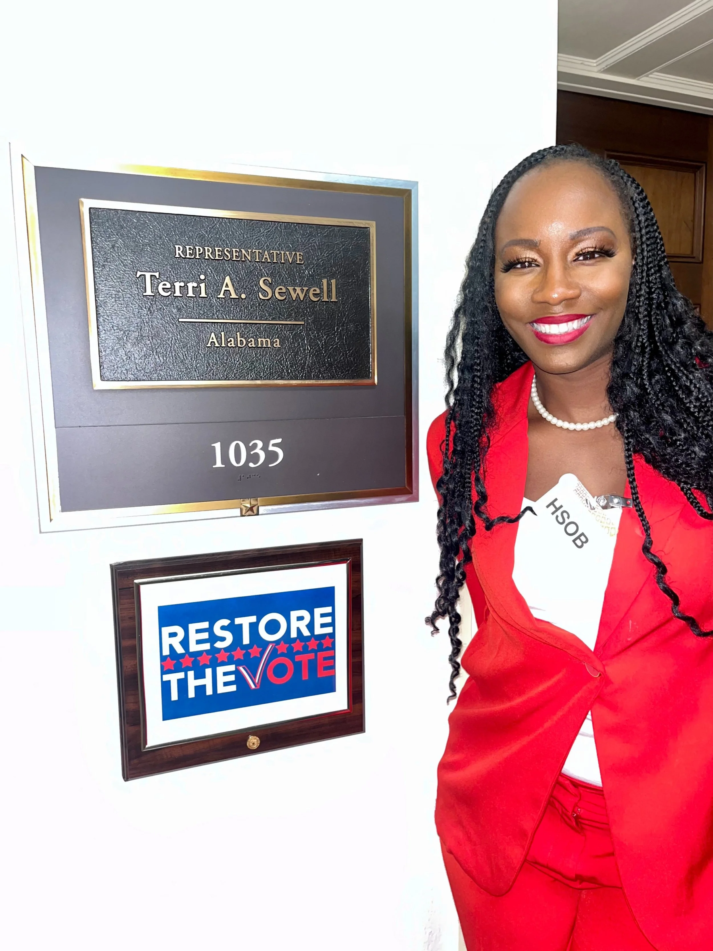 A woman in a red suit standing outside a door with a nameplate that reads 'Representative Terri A. Sewell, Alabama, 1035.' She is smiling and wearing a pearl necklace with a badge labeled 'HSOB.'