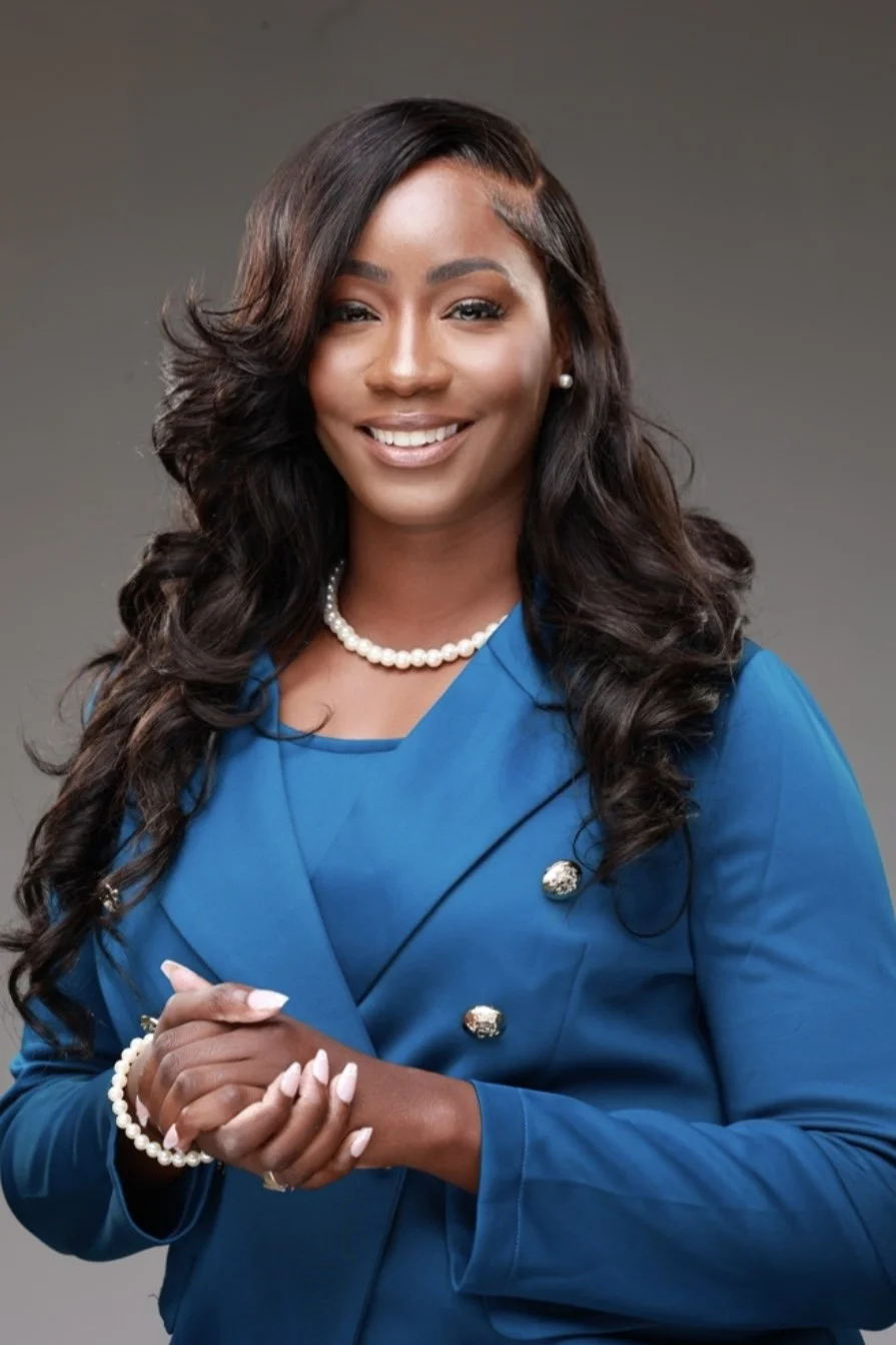 A professional woman with dark wavy hair, wearing a blue blazer, pearl necklace, pearl earrings, and a pearl bracelet, smiling and posing with her hands clasped in front of her against a plain gray background.
