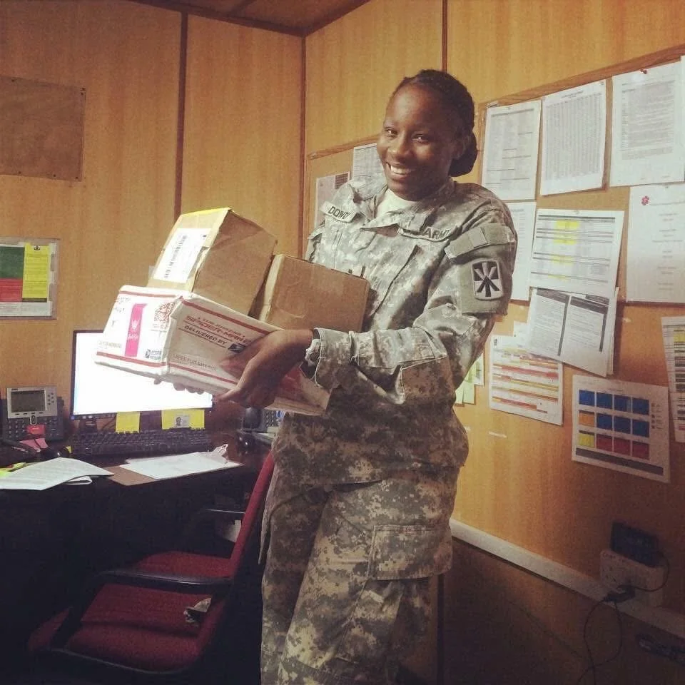 A woman in a military uniform holding a stack of boxes inside an office with charts and papers on the walls.