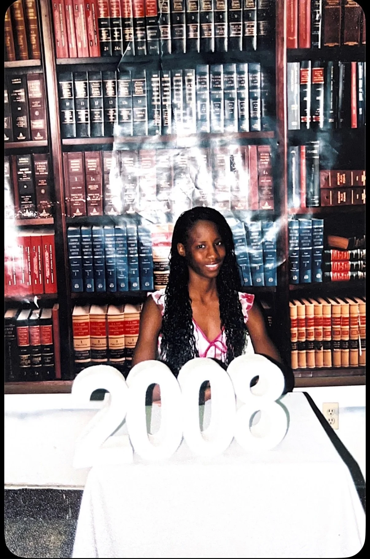 A woman sitting at a table with large white numbers reading '2008' in front of her, in front of a background of bookshelves filled with legal books.