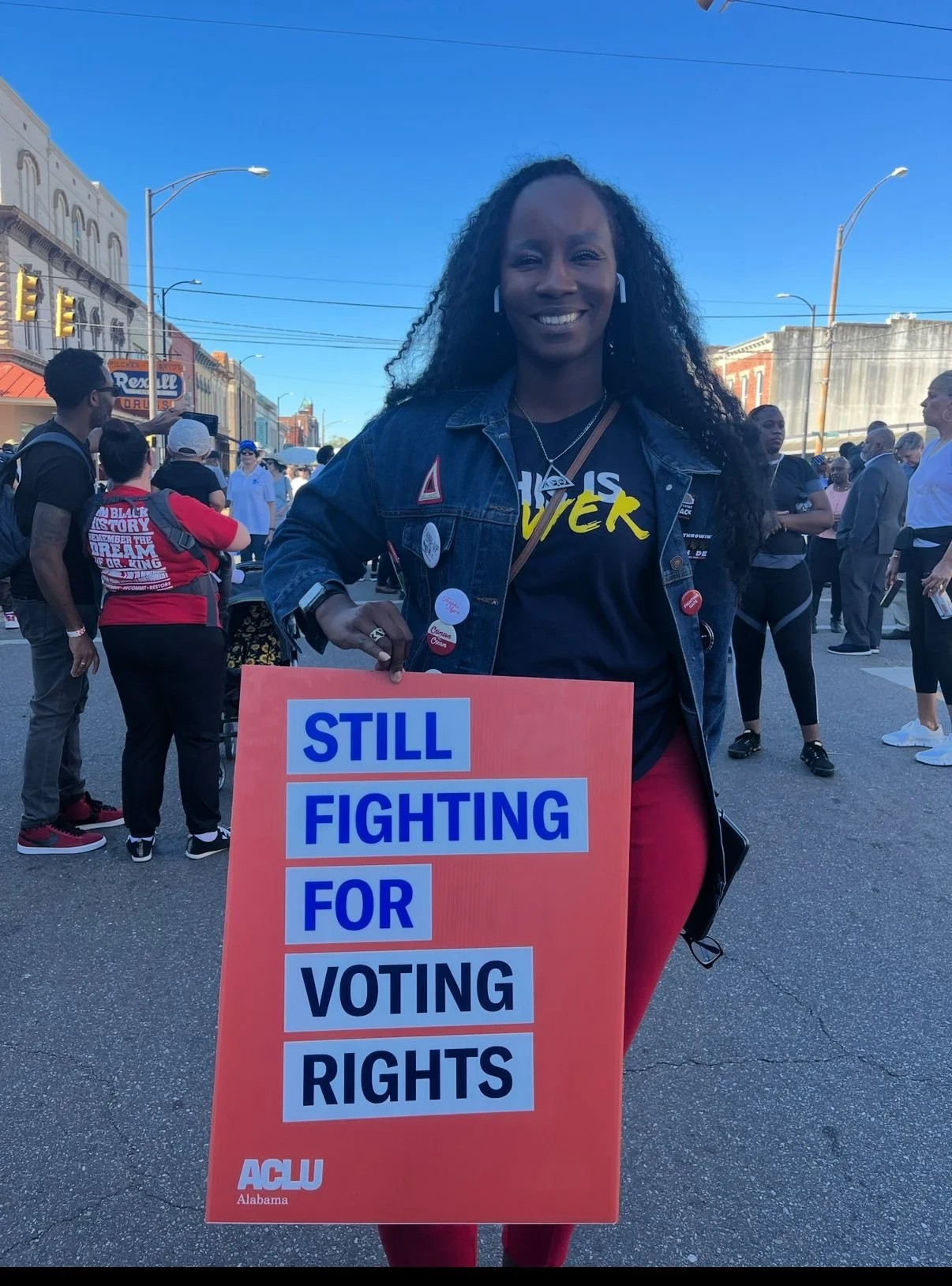 A woman at a rally holding a sign that reads, 'Still fighting for voting rights,' smiling and standing among other protesters on a city street during daytime.