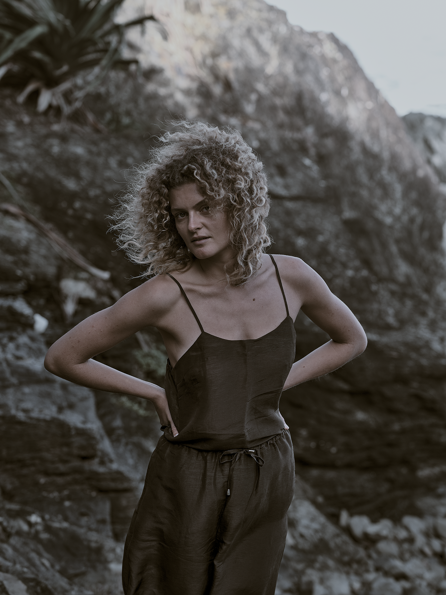 Woman with curly hair in a black top and pants standing outdoors in front of rocks and foliage.