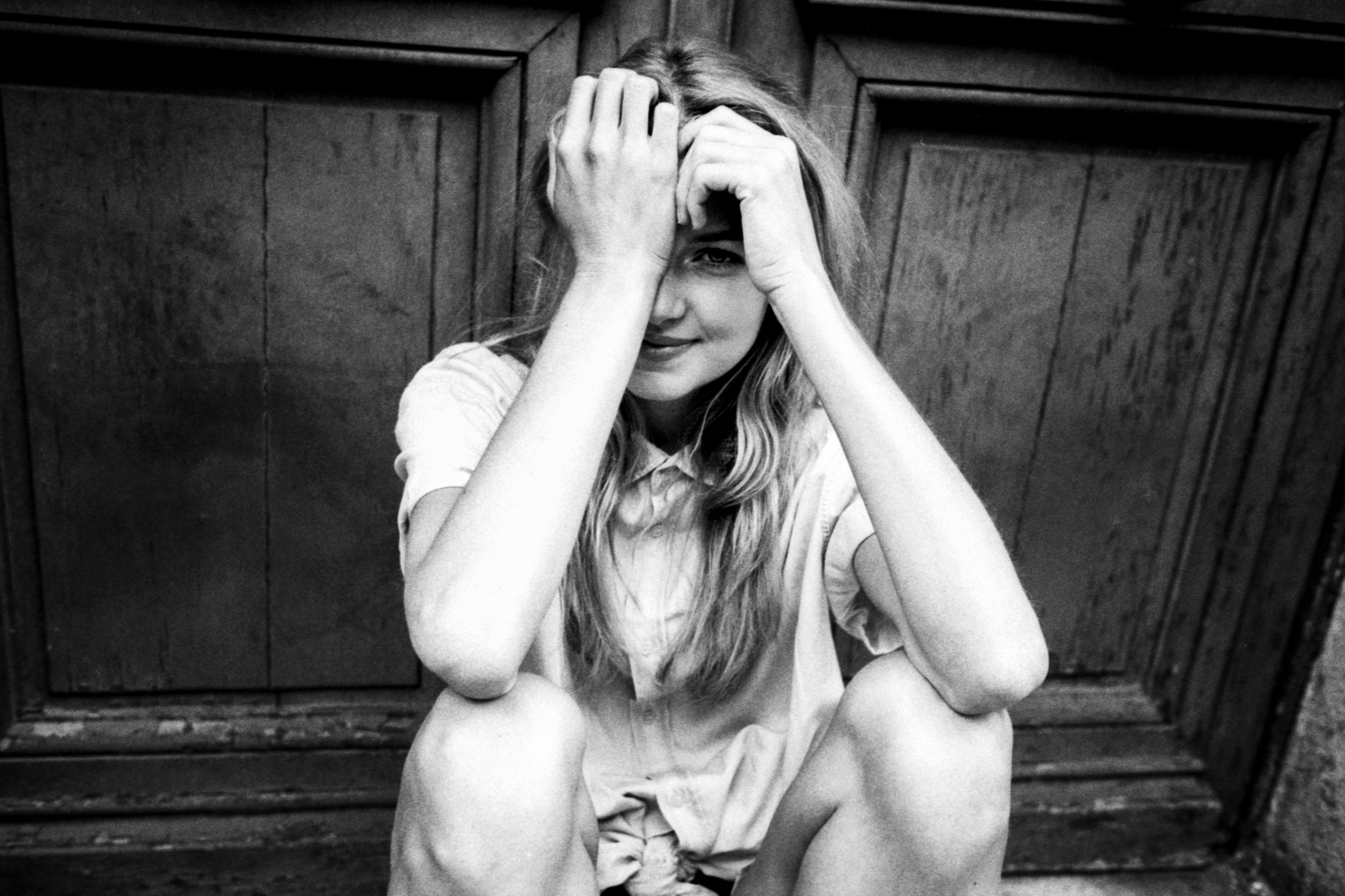 A woman with long hair is sitting on the ground with her back against a wooden wall. She covers part of her face with her hands and appears to be smiling or smirking.