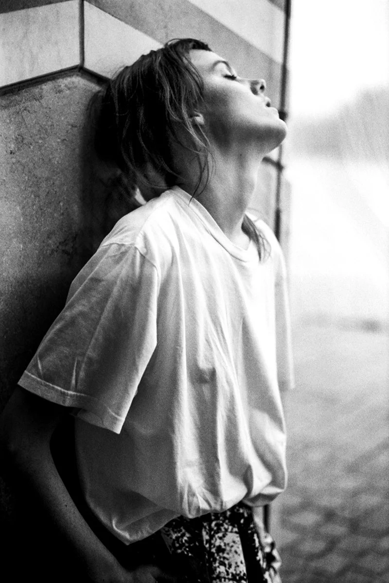 Black and white photo of a woman with short hair leaning against a tiled wall, eyes closed, with a contemplative expression.