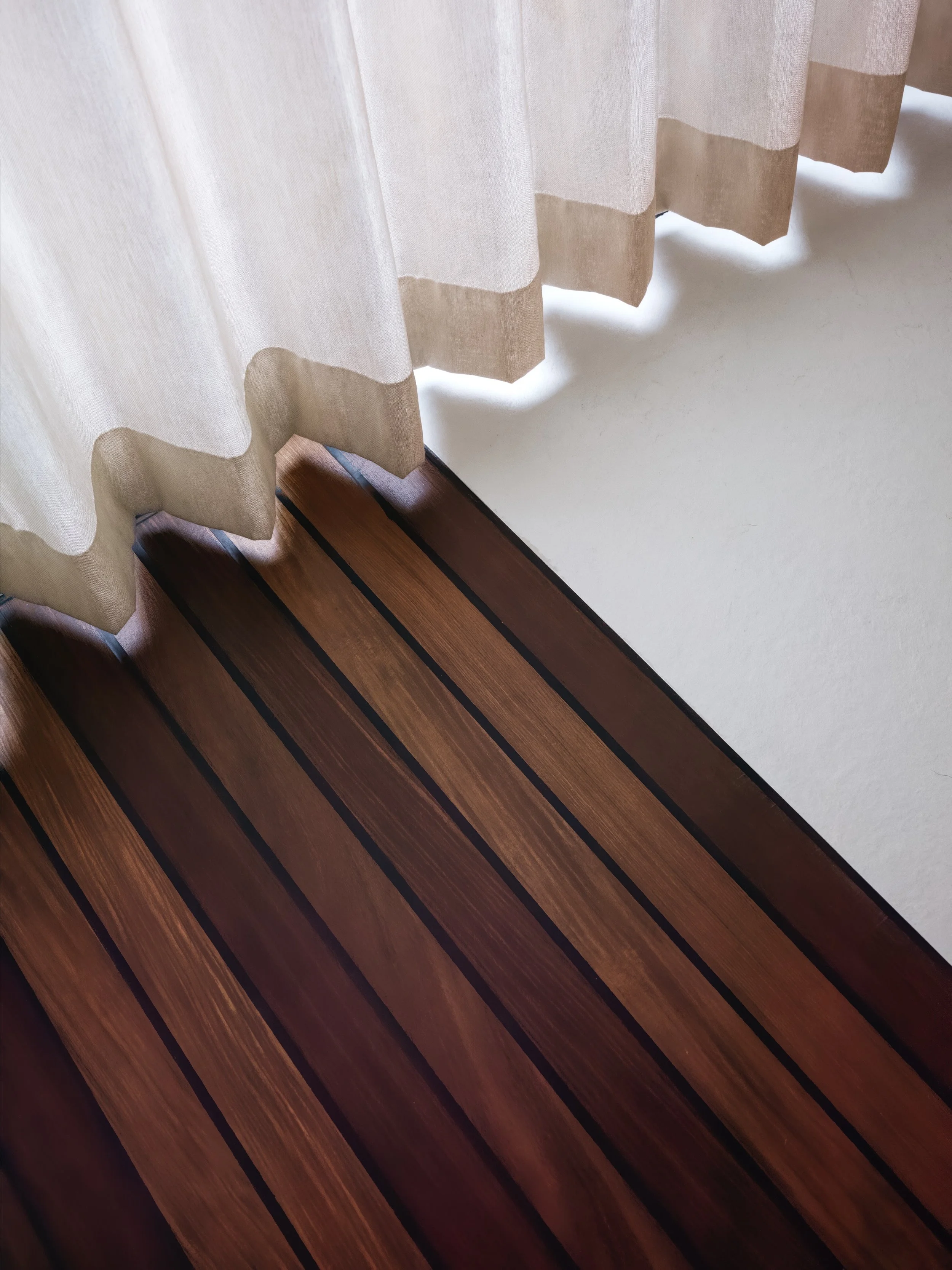 View of a dark wood floor next to a white wall with white curtains with beige trim hanging over it.