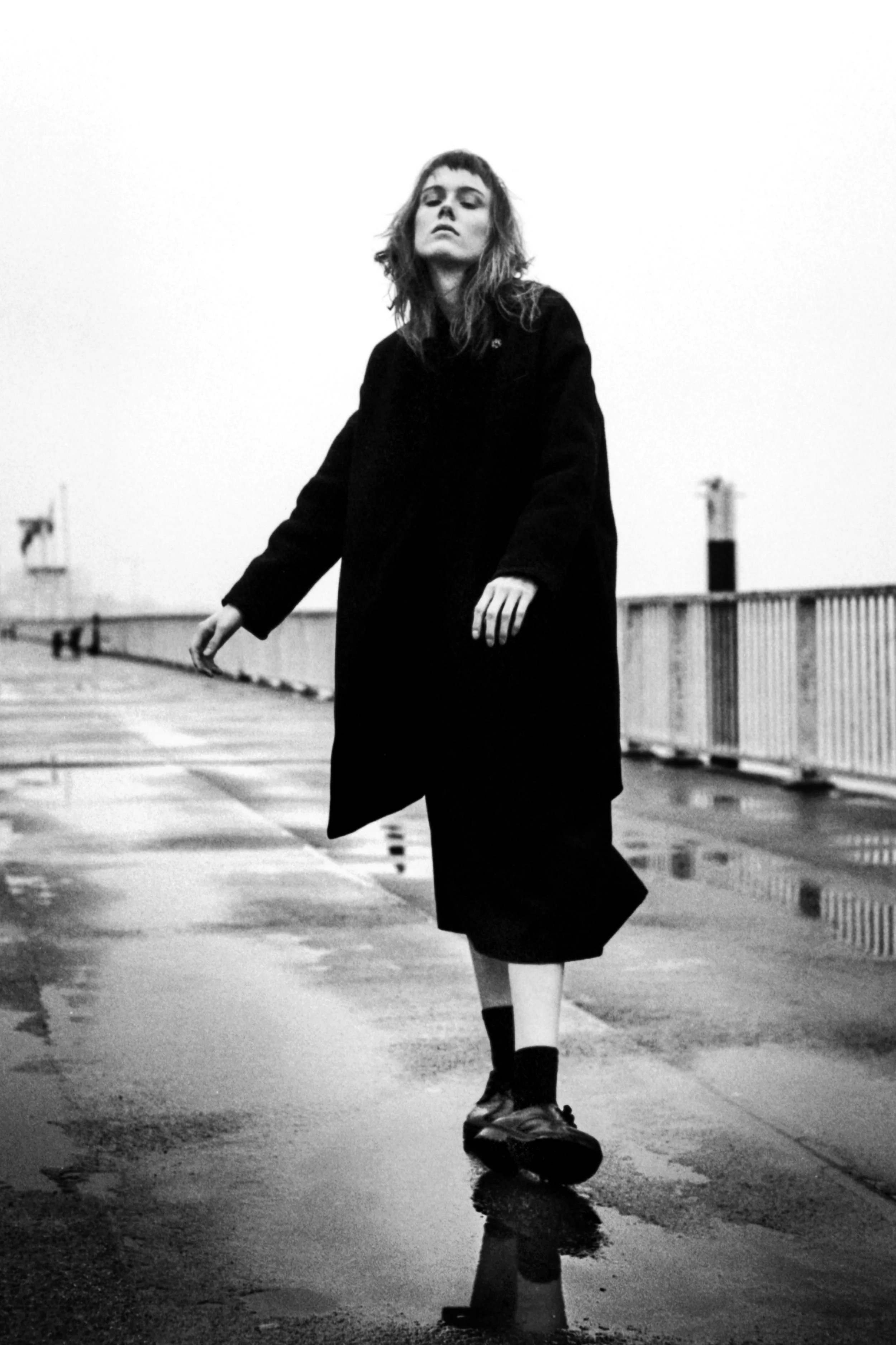 A young woman with shoulder-length hair and bangs, wearing a dark coat and skirt, is standing on a wet surface, possibly a street or pier, with a railing and some indistinct structures in the background. She is looking down at a skateboard she is sta