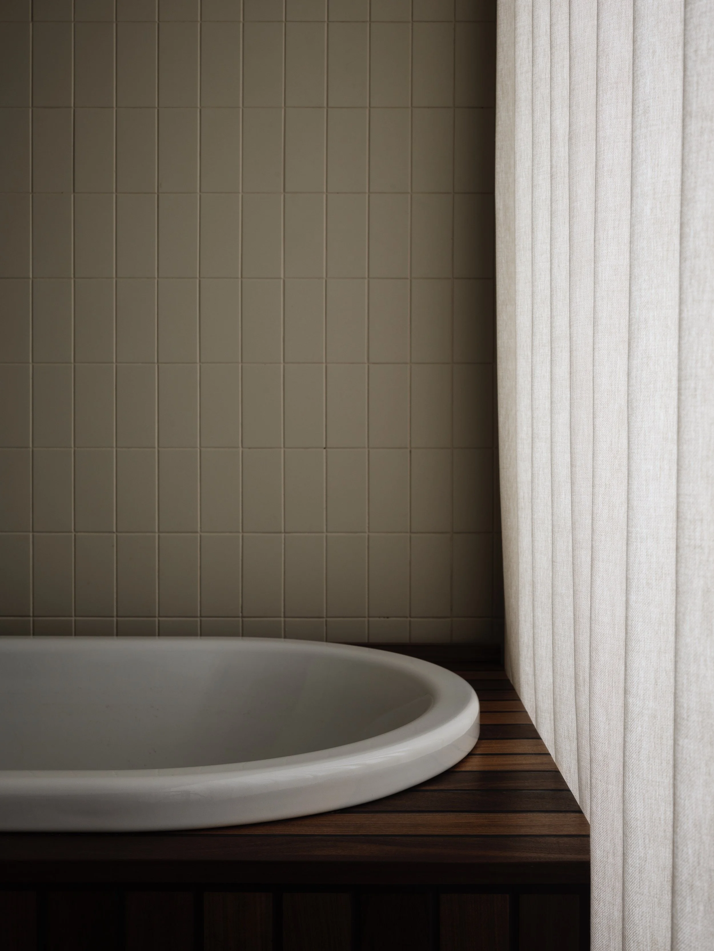 Close-up of a gray oval bathtub on a wooden platform against a tiled wall with beige curtains on the side