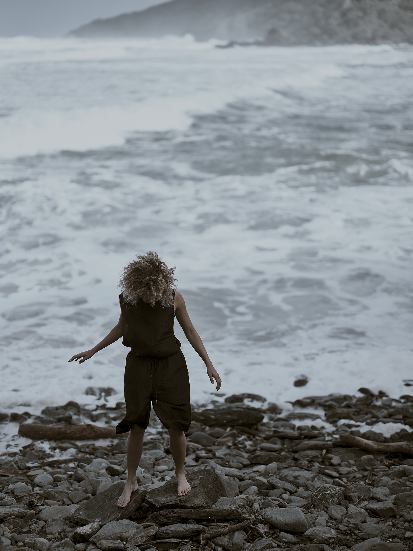 Person standing barefoot on rocks at the beach, facing the ocean, with waves crashing and distant foggy hills in the background.