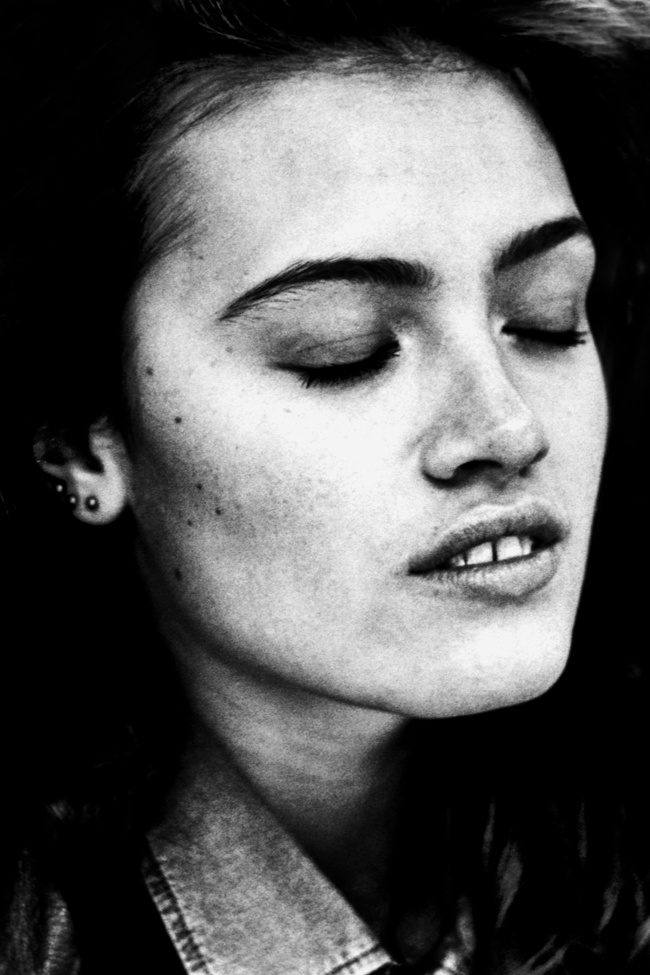 Black and white close-up photo of a woman's face with her eyes closed, showing her eyebrows, nose, lips, earrings, and part of her neckline.