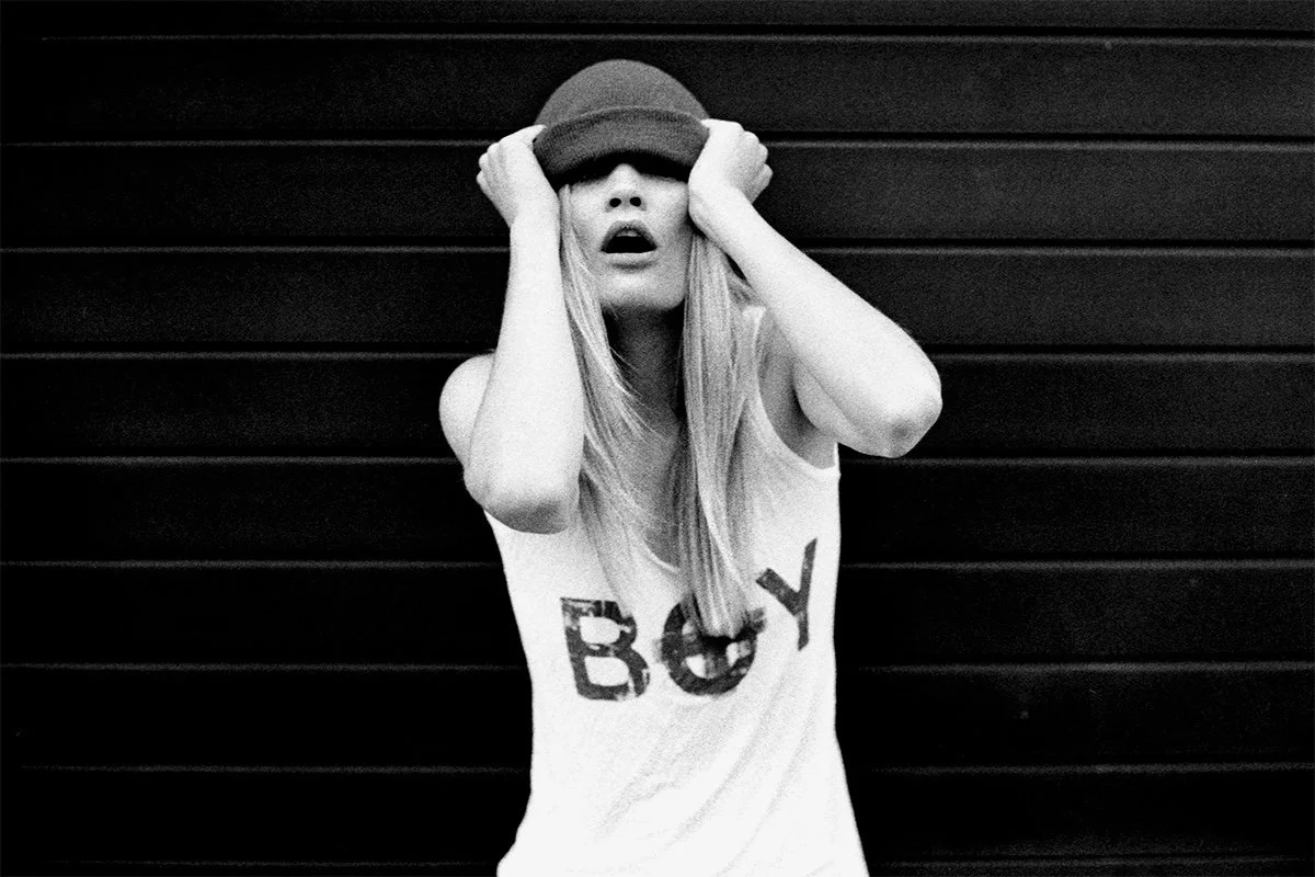 A woman with long blonde hair covering her eyes with a hat and her hands, standing against a dark horizontal panel background.