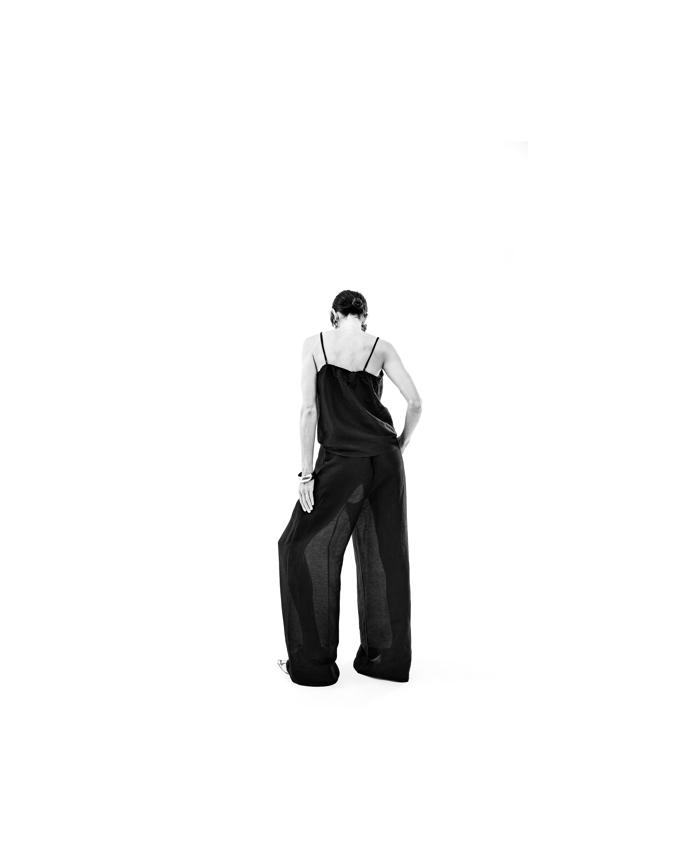 A woman with short hair standing with her back to the camera, wearing a loose black satin top and wide-leg black pants, in a minimalistic setting with a plain white background.