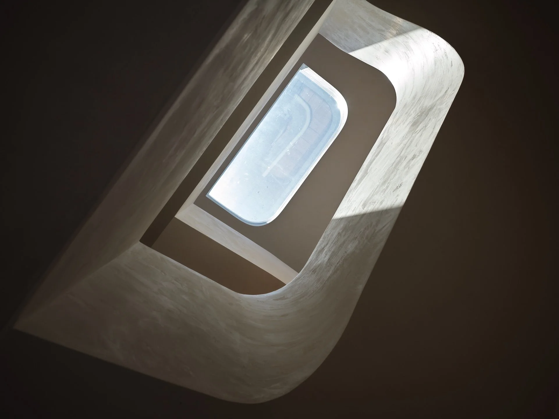 Looking up through a square opening in a spiral staircase with a window at the top, sunlight casting shadows.