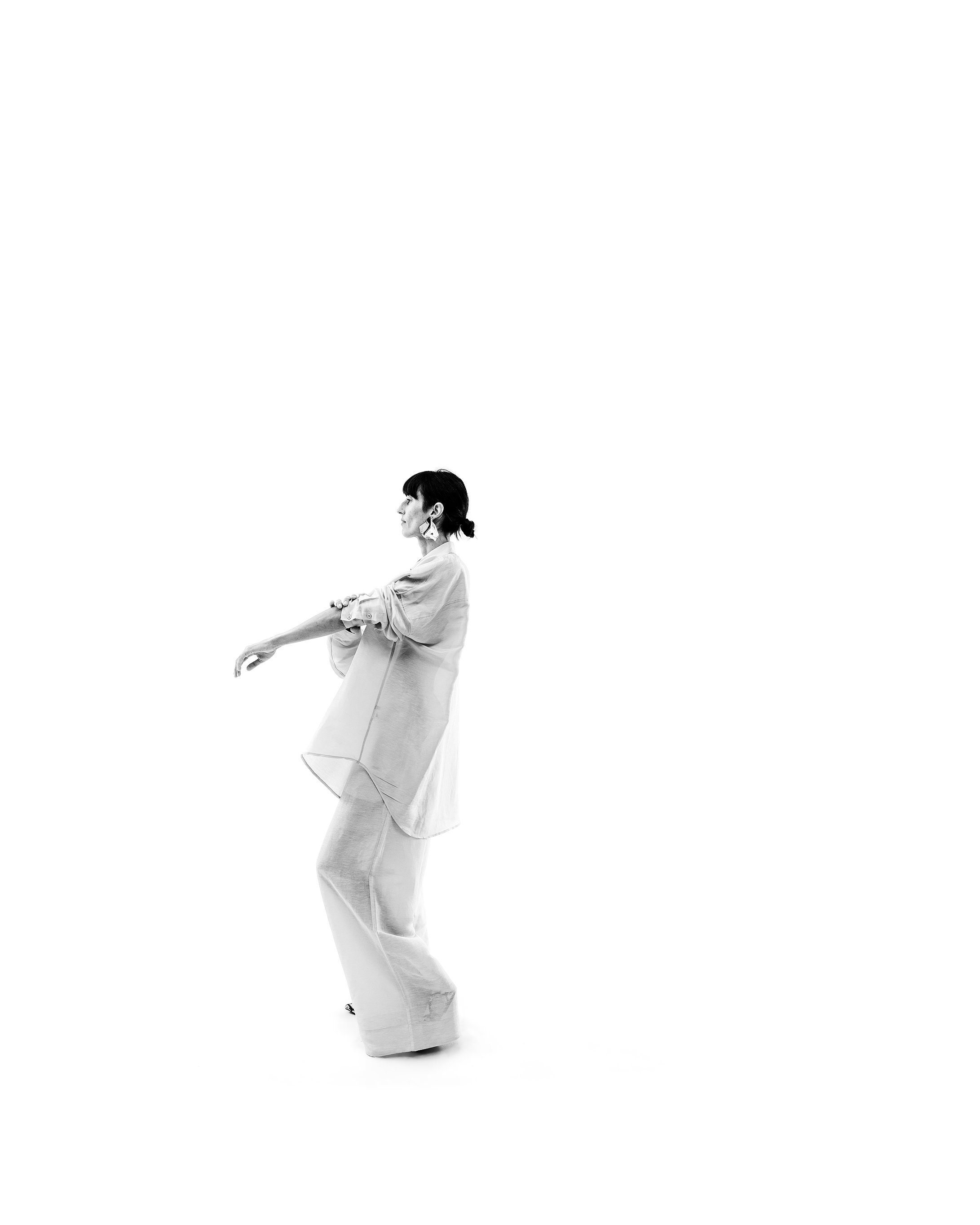 A woman dancing against a white background, dressed in a loose, light-colored outfit, with short dark hair and earrings, captured in a black and white photograph.