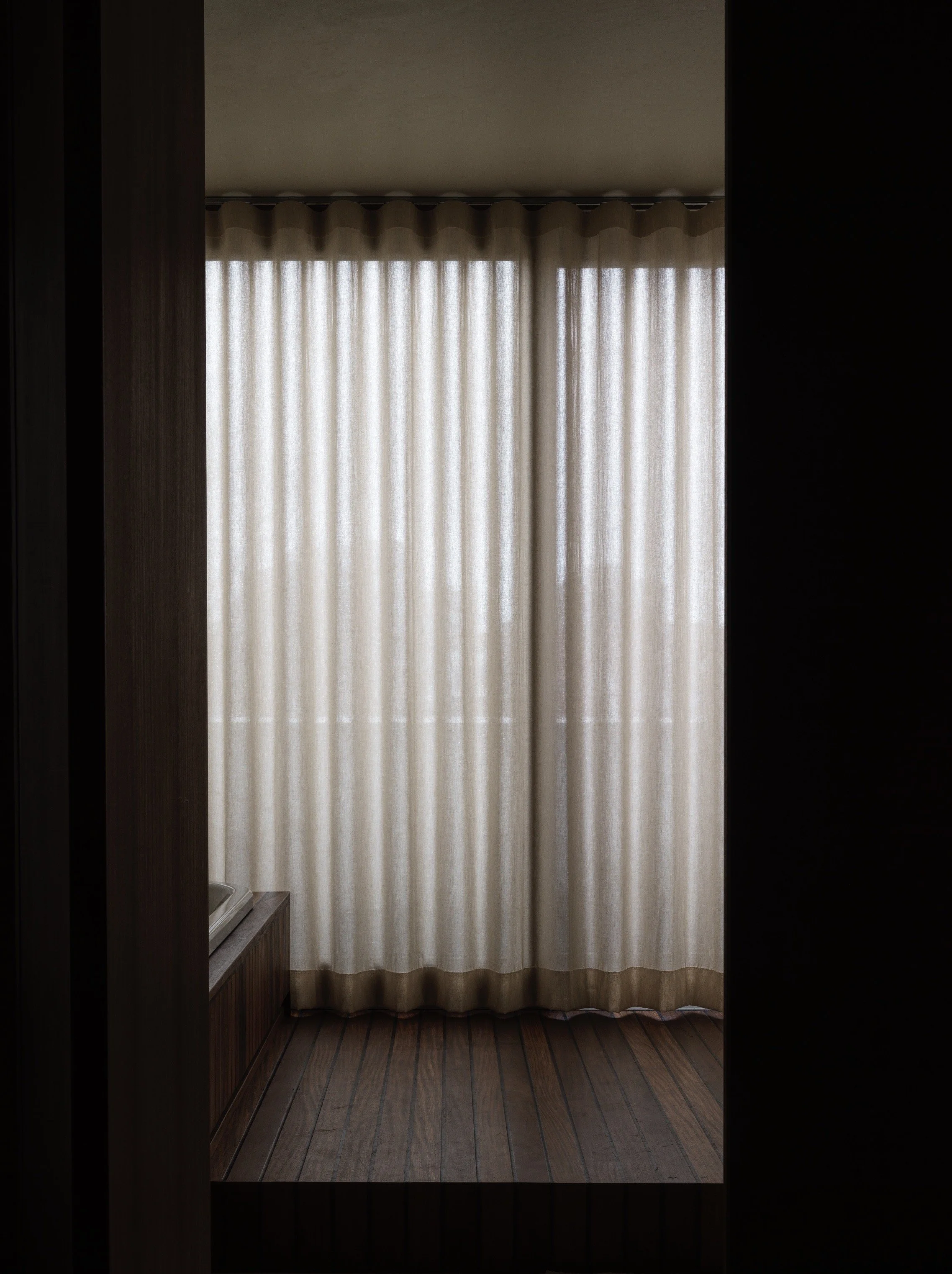 A window with beige curtains drawn closed, framed by dark walls and wood flooring.