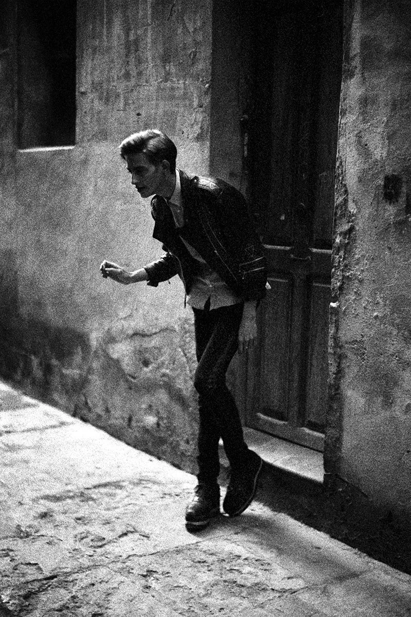 A young man wearing a leather jacket, white shirt, and dark pants, standing on a cobblestone street near a doorway in an old, textured wall, captured in black and white.