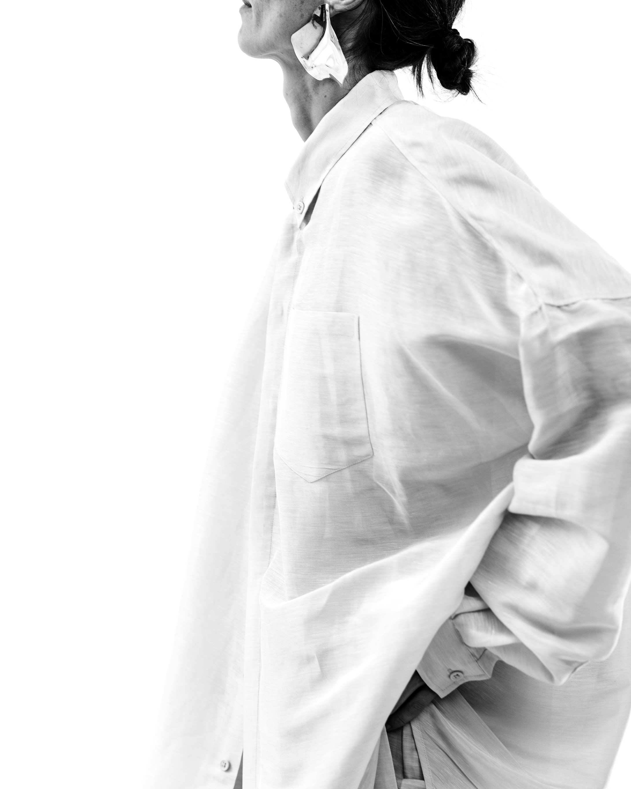 A woman in a white oversized shirt, with her hand in her pocket, wearing reflective sunglasses and a bun hairstyle, against a plain white background.